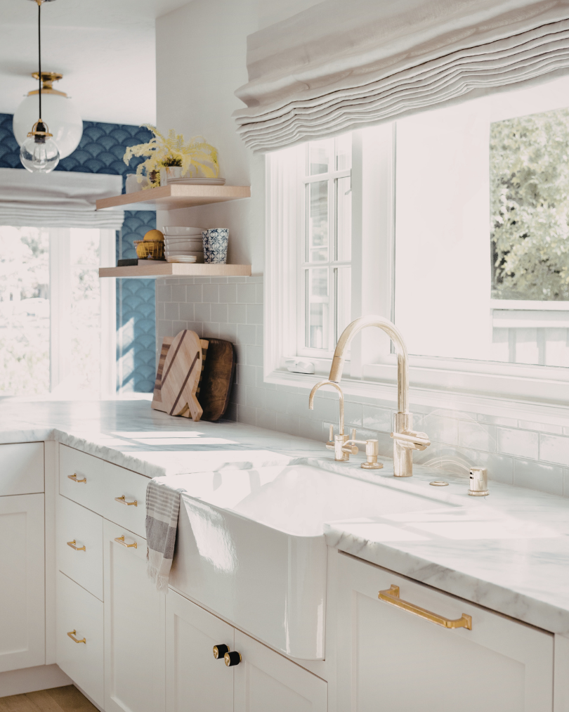 Bright kitchen with white cabinetry, marble countertop, and a white farmhouse sink with a high-arc gold faucet. Open shelves displaying dishes and decor, large windows with white blinds, and sunlight filling the space.
