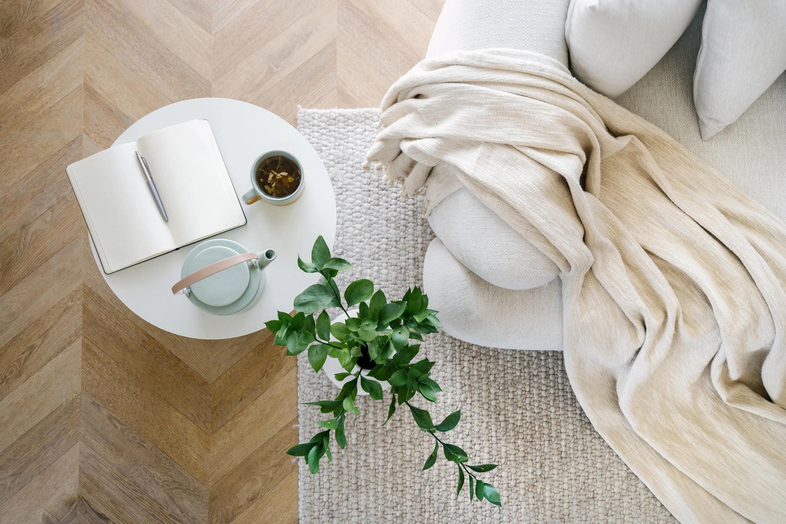 Top-down view of a cozy living room corner with a light-colored sofa, a beige throw blanket, a small round white side table holding an open notebook, pen, mug of tea, teapot, and a green potted plant, on a light textured rug and hardwood flooring.