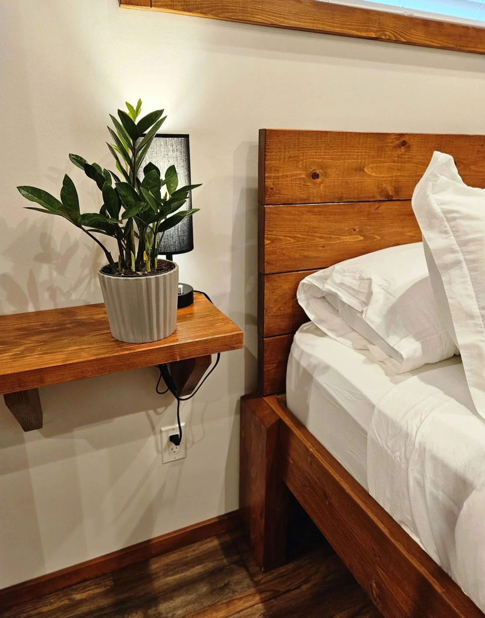 A bedroom with a wooden bed frame, white bed linens, a bedside table with a potted plant, a black lamp, a power outlet with a plug, and wooden flooring.