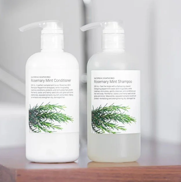 Two white bottles of Rosemary Mint shampoo and conditioner on a wooden surface, with a gray background.