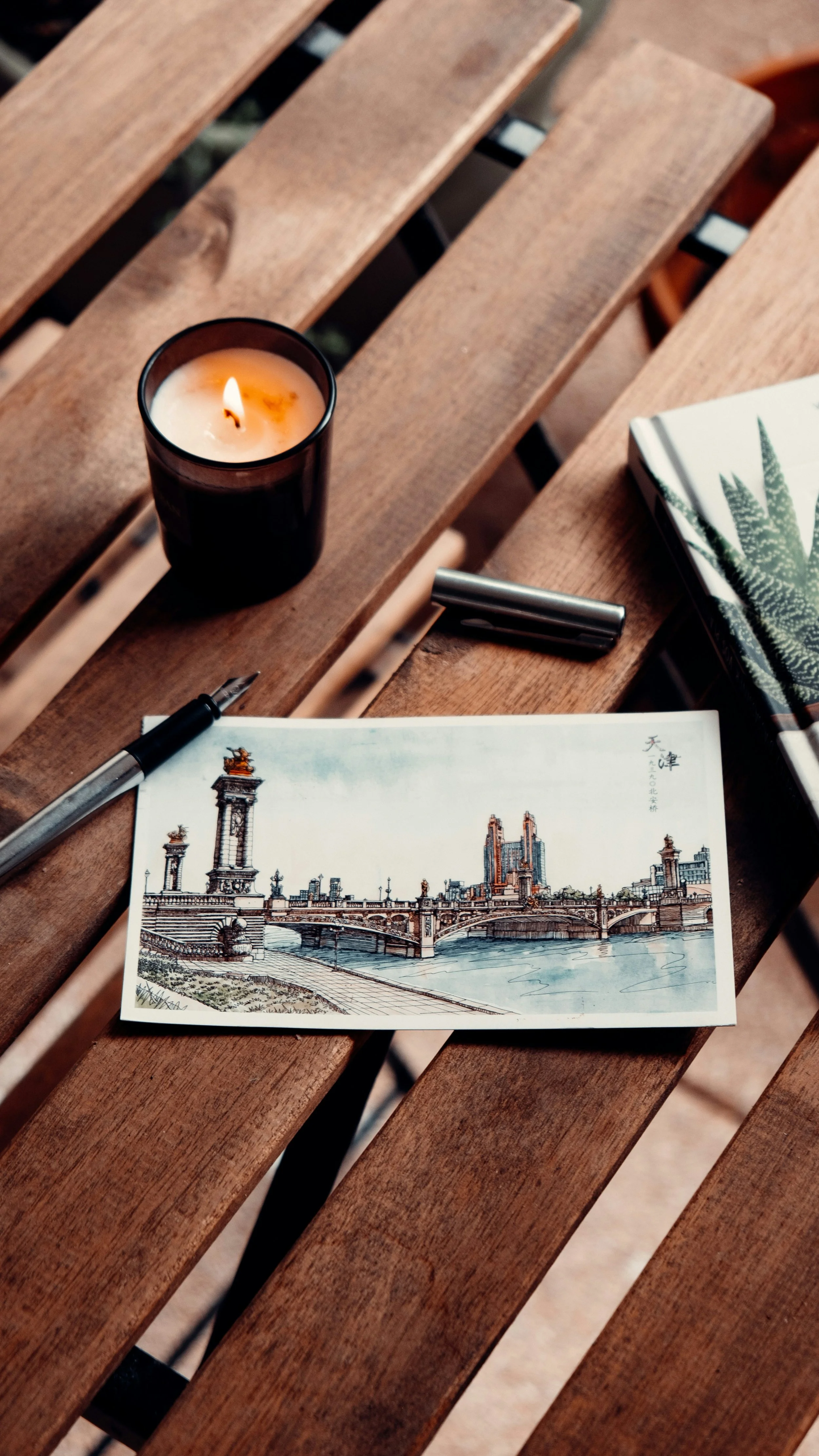 A candle in a black holder, a postcard of a cityscape with a bridge and tall buildings, a fountain pen, a closed book with a green plant illustration, and a small container are arranged on a wooden table.
