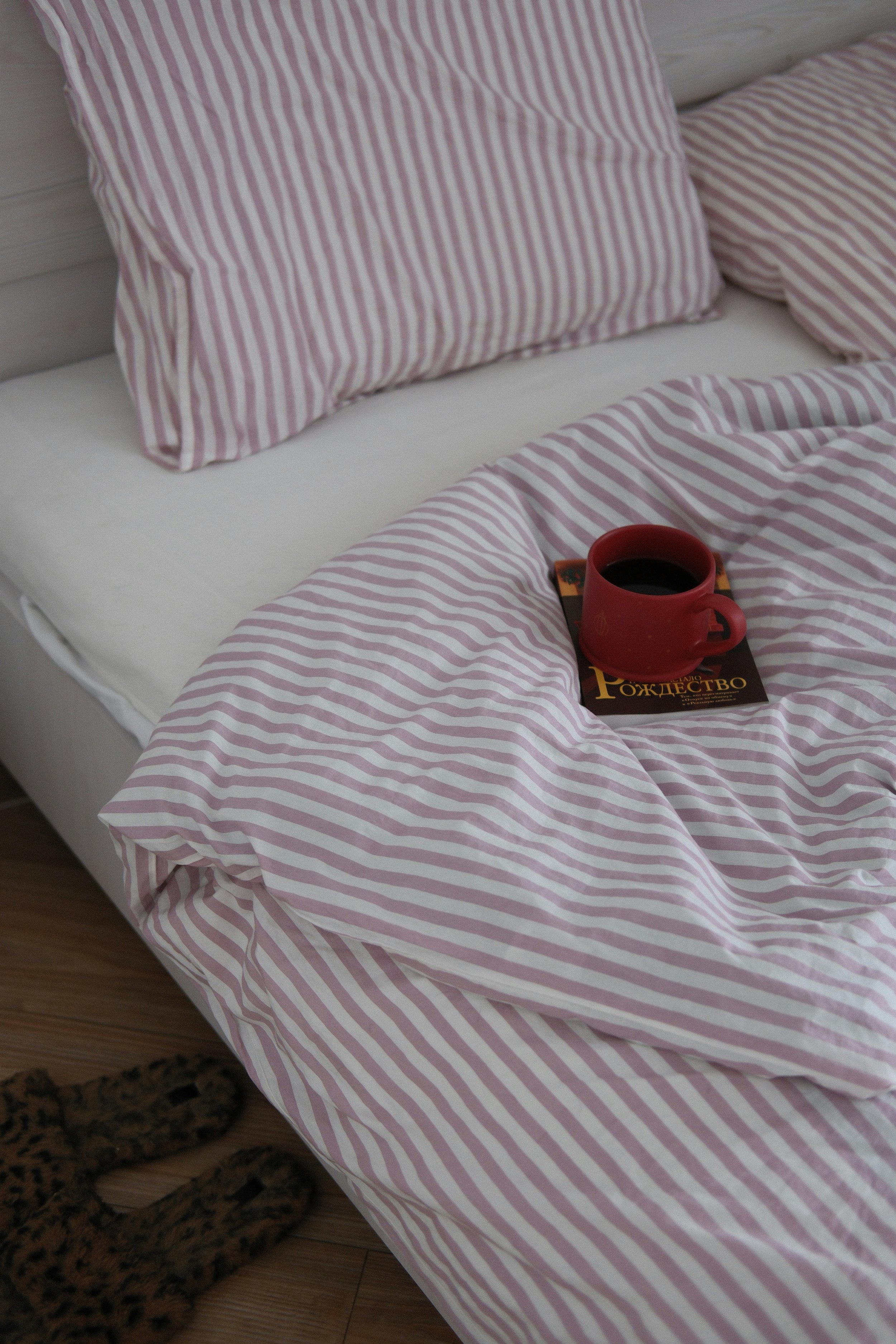 A bed with pink and white striped sheets and matching pillows. On the bed, there is a red mug of coffee on top of a book written in Cyrillic script. There is a leopard print blanket at the foot of the bed, and the floor appears to be wooden.