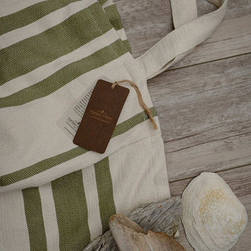 A green and white striped tote bag with a tag labeled "Tofino Towel" lying on a wooden surface near a piece of driftwood and a seashell.