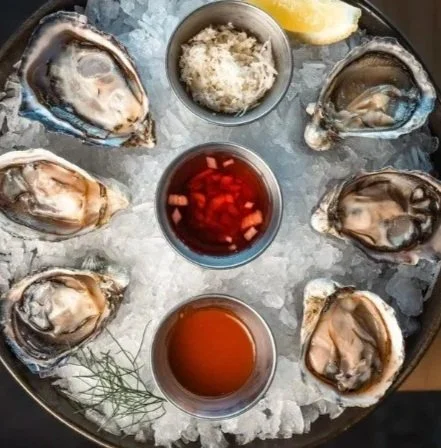 Oyster shellfish platter with fresh oysters on ice, lemon wedge, grated horseradish, cocktail sauce, mignonette sauce, and hot sauce.