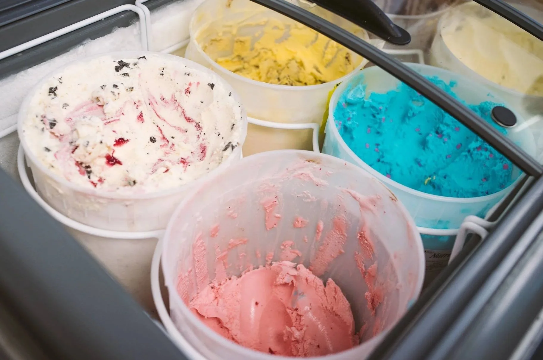 Various flavors of ice cream in tubs inside a freezer, including strawberry, blue raspberry, vanilla, and cookies and cream.