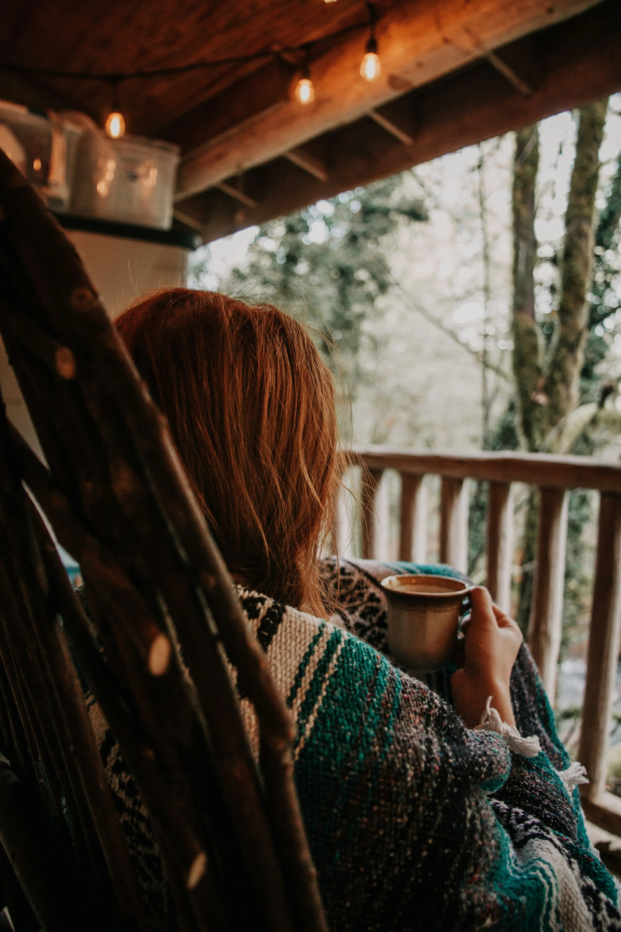 Person with reddish hair sitting on a porch or balcony, holding a mug, overlooking a forested area with trees, under a wooden roof with hanging lights.