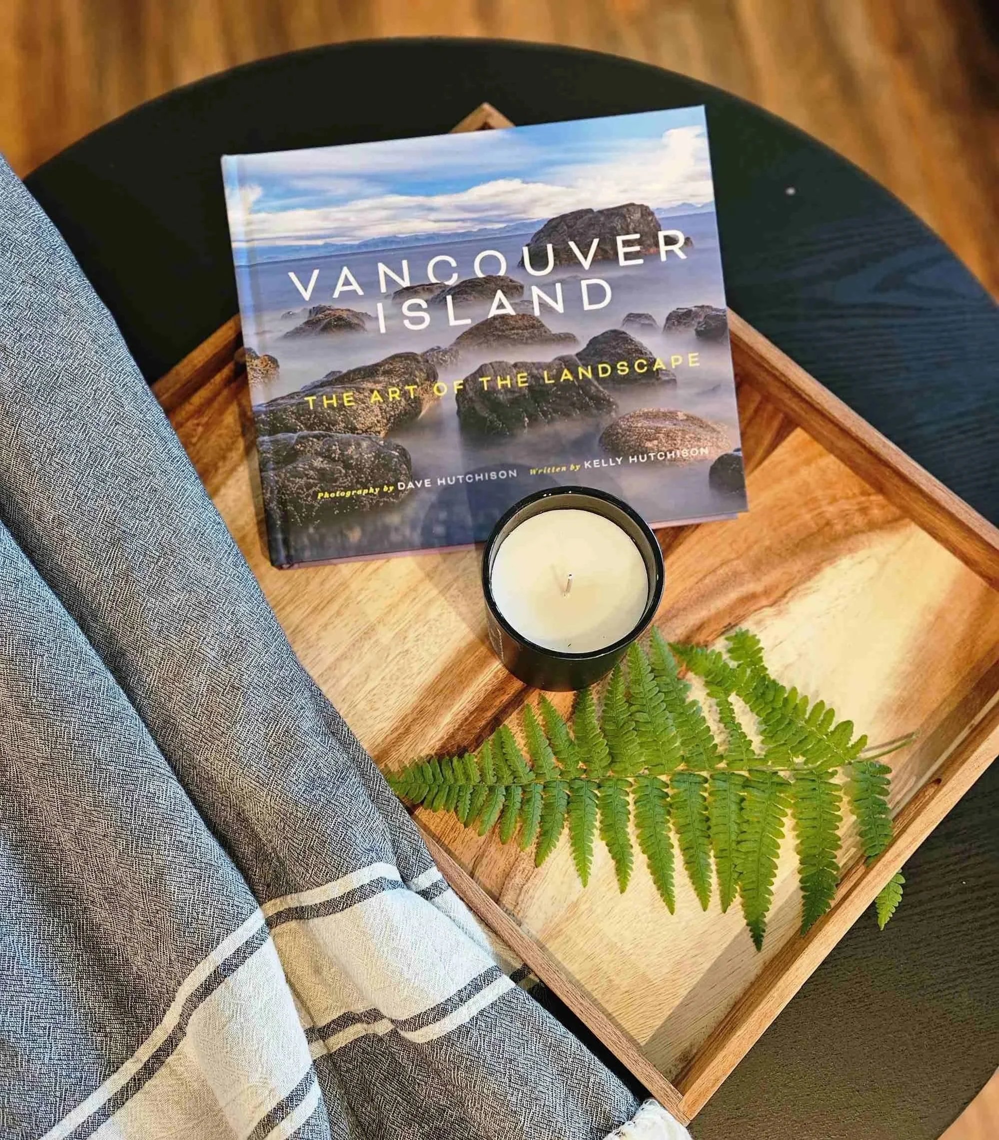 A book titled 'Vancouver Island: The Art of the Landscape' on a wooden table, with a green fern and a white candle in a black holder nearby. A person wearing gray checkered pants and a white and black striped towel is sitting next to the table.