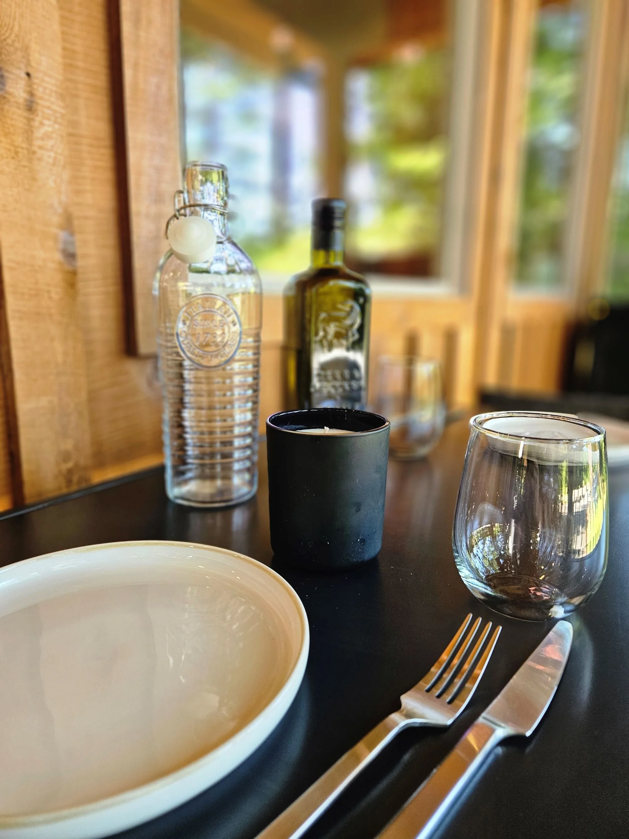A dining table set with a white bowl, a black candle holder with a white candle, a clear glass, a water bottle, and a wine bottle with a blurred wooden window in the background.