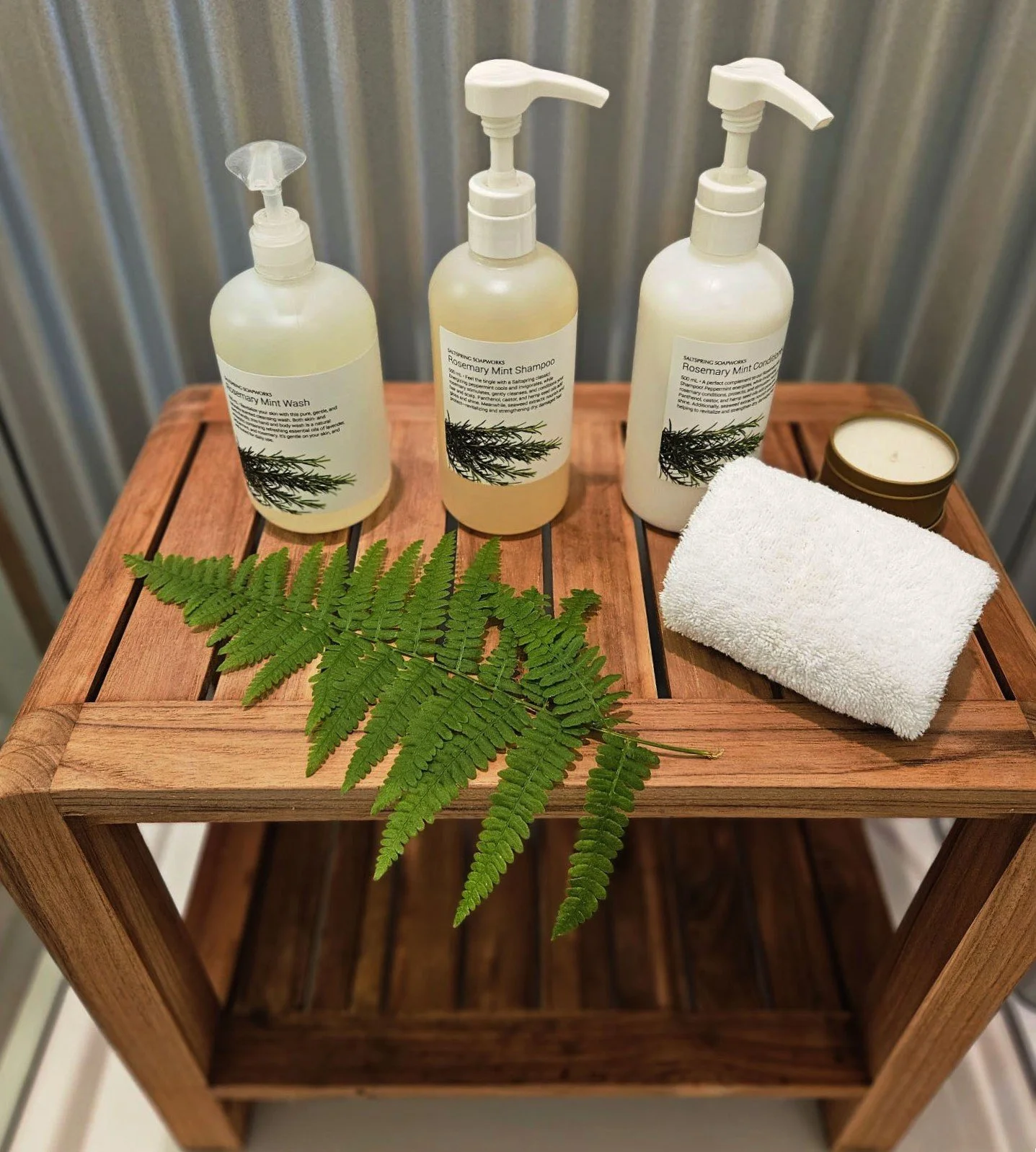 A wooden shelf with three bottles of rosemary mint soap, a green fern leaf, a white towel, a candle, and labels on the bottles.