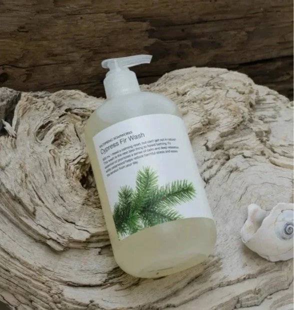A bottle of cypress fir wash with a pump dispenser resting on a piece of driftwood next to a seashell.