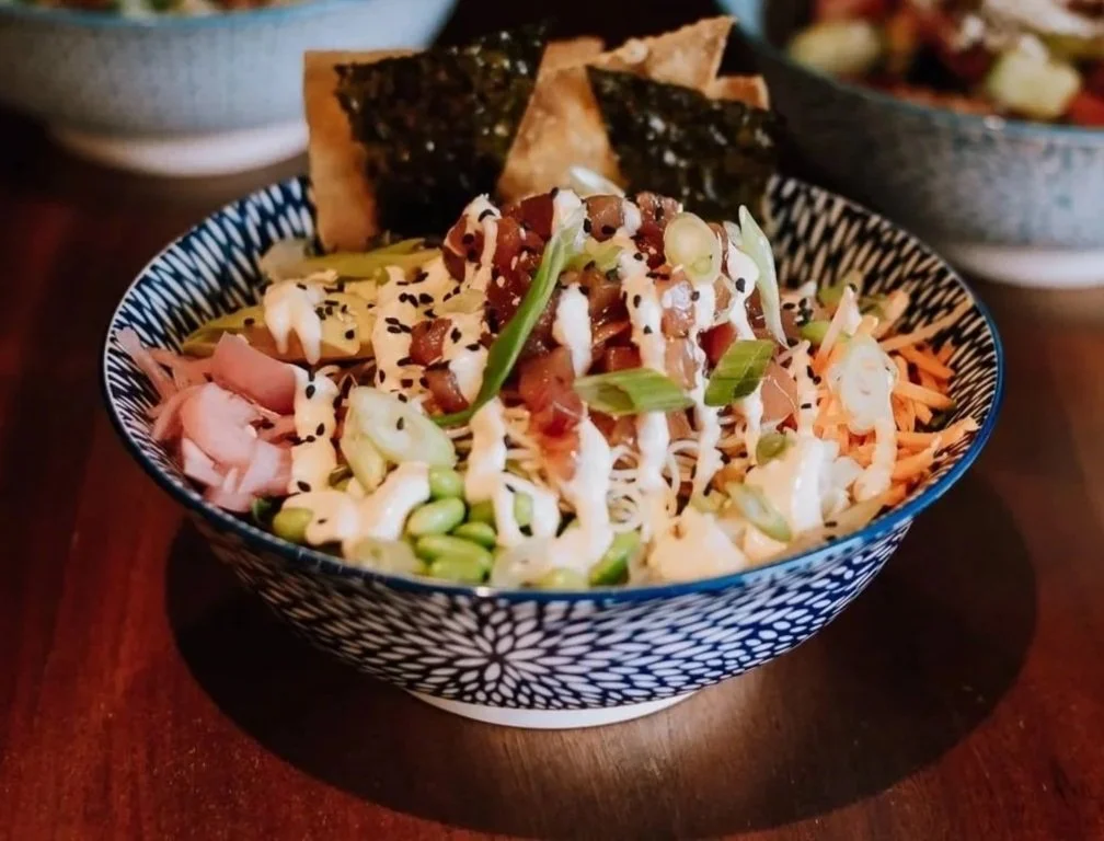 Bowl of Asian-inspired salad salad topped with sliced green onions, shredded carrots, shredded chicken, and drizzled with mayonnaise or dressing, with seaweed crackers on top. In the background, there are two more bowls, one with a salad and the other with a different dish.