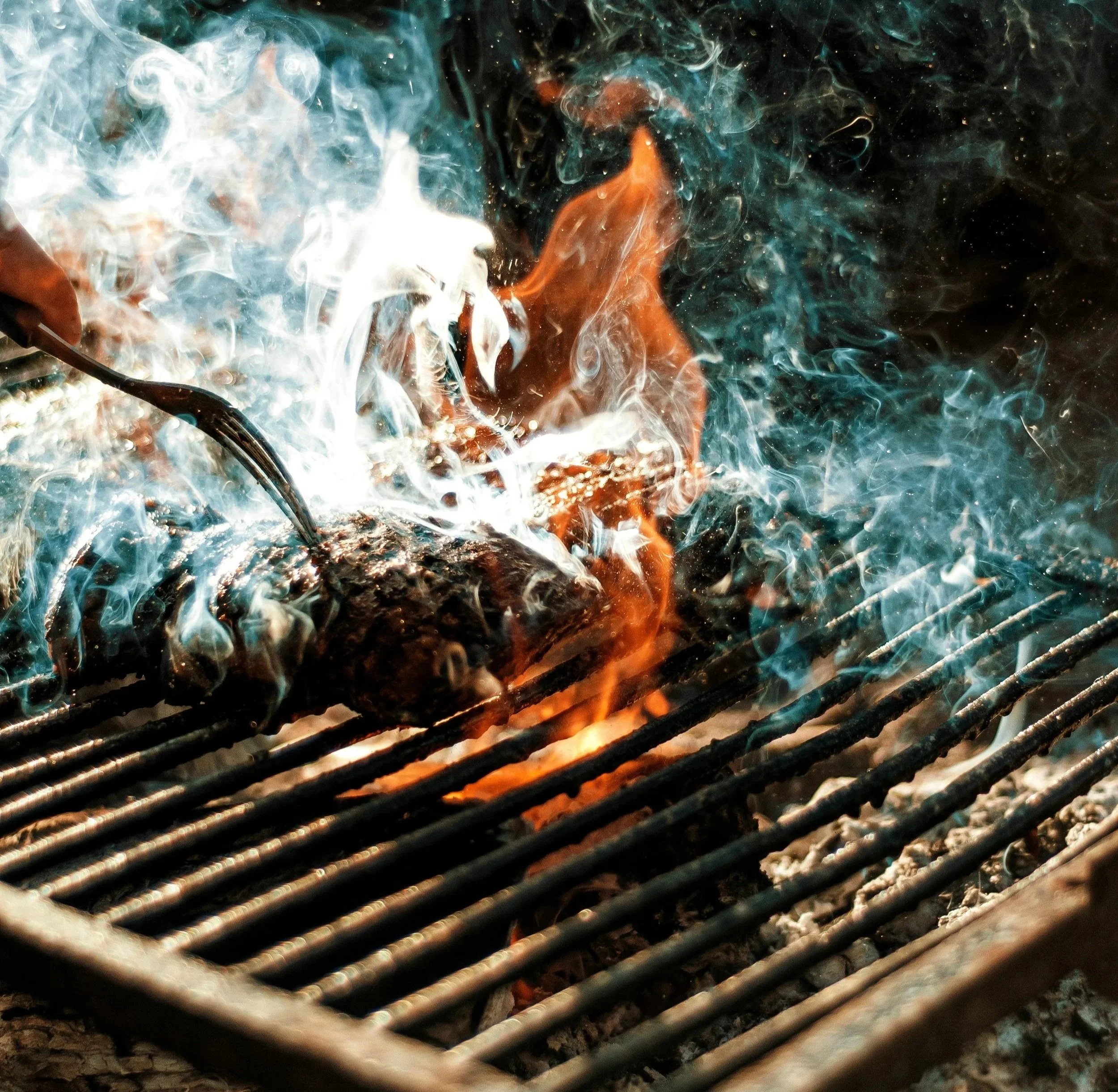 Close-up image of a grill with glowing hot charcoal and orange flames visible beneath metal grates, with smoke rising above.