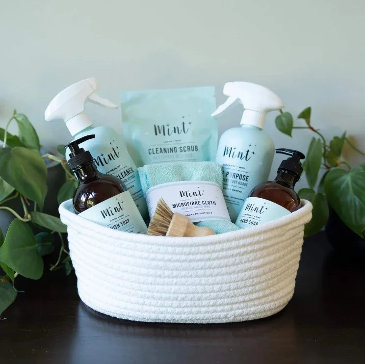 A white, textured oval basket containing various eco-friendly cleaning and personal care products, including spray bottles, liquid soap, a microfiber cloth, a small brush, and a cleaning scrub, surrounded by green leafy plants.
