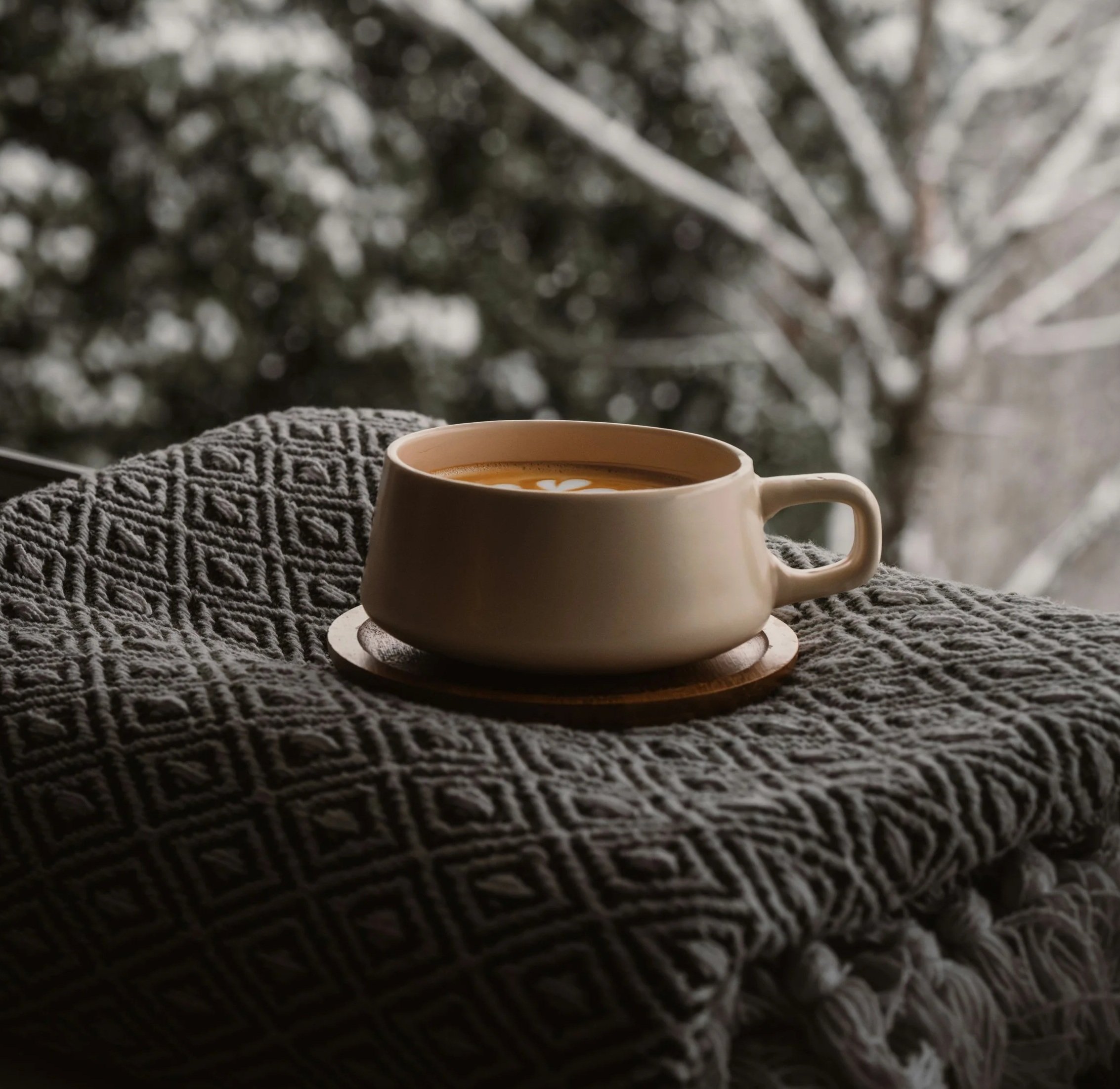 A close-up of a white ceramic mug filled with coffee with latte art on top, placed on a textured gray knitted blanket near a window with a snowy scene outside.