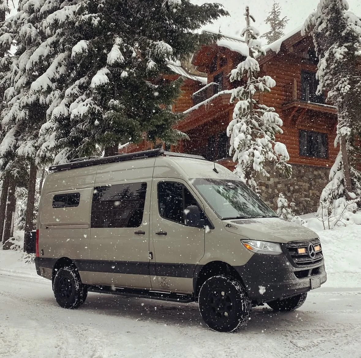 A beige Mercedes-Benz Sprinter van parked on a snowy road, with a wooden house and snow-covered trees in the background during a snowfall.