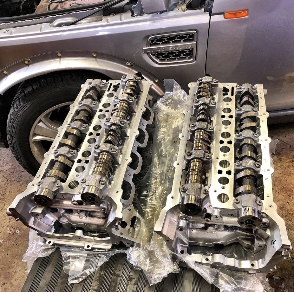 Two engine cylinder heads and camshafts on a workbench in front of a gray Land Rover vehicle.