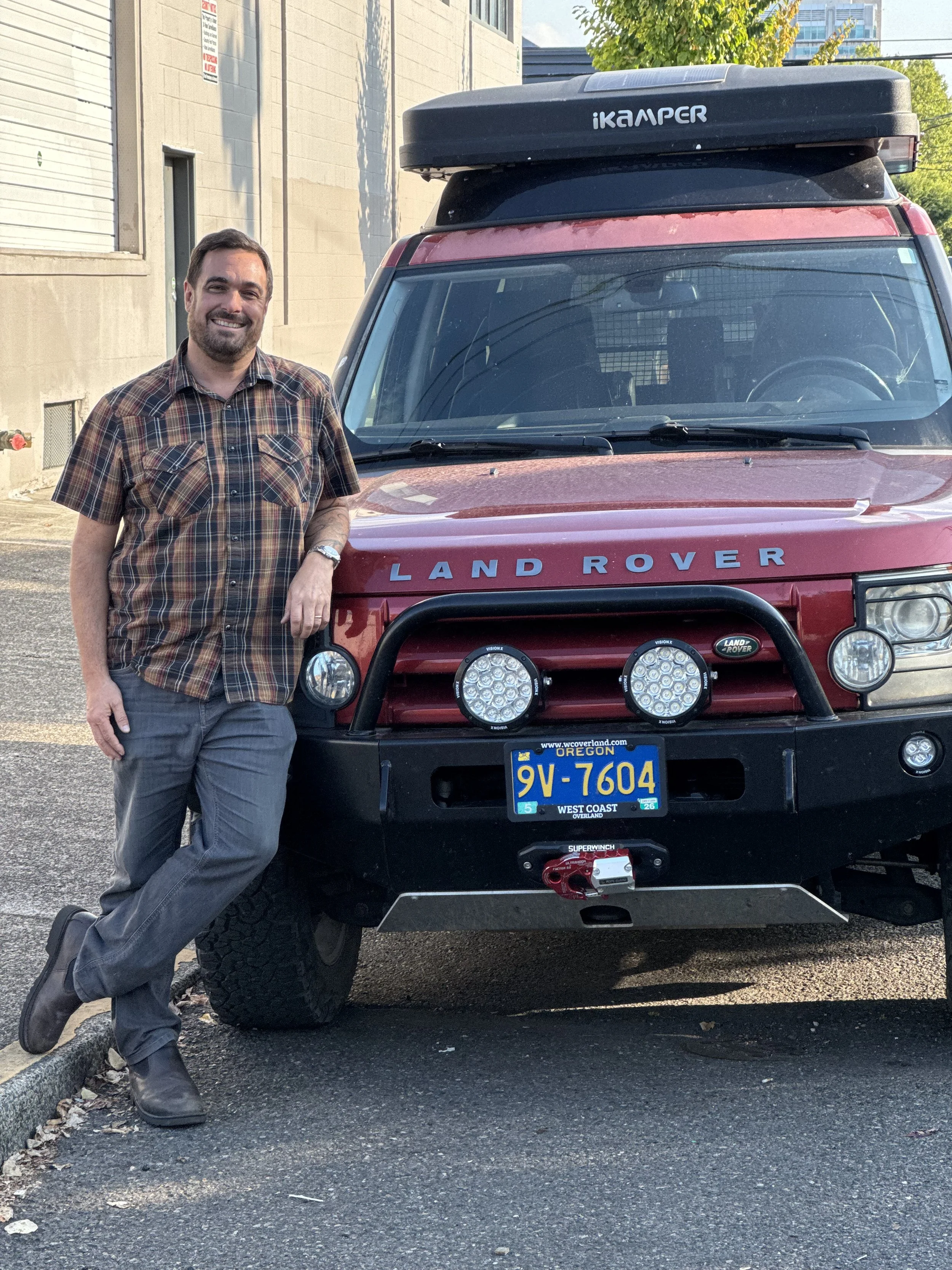 A man with a beard, wearing a plaid shirt and gray pants, smiling and leaning against a red Land Rover vehicle in a parking lot.