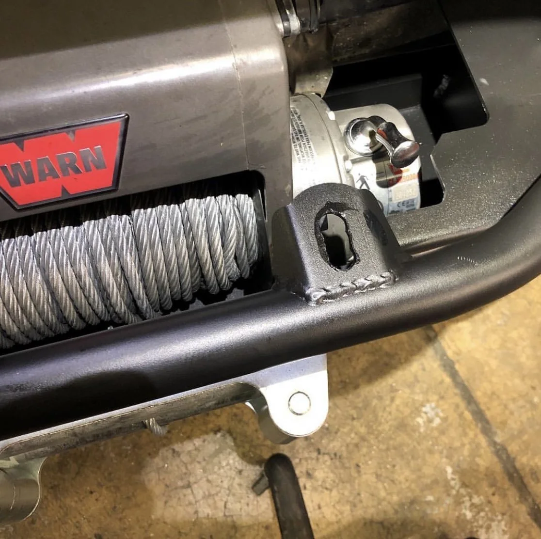 Close-up of a winch with a metal cable, part of a mechanical or industrial setup, with a red and black 'WARN' label, on a metallic surface with a black frame.