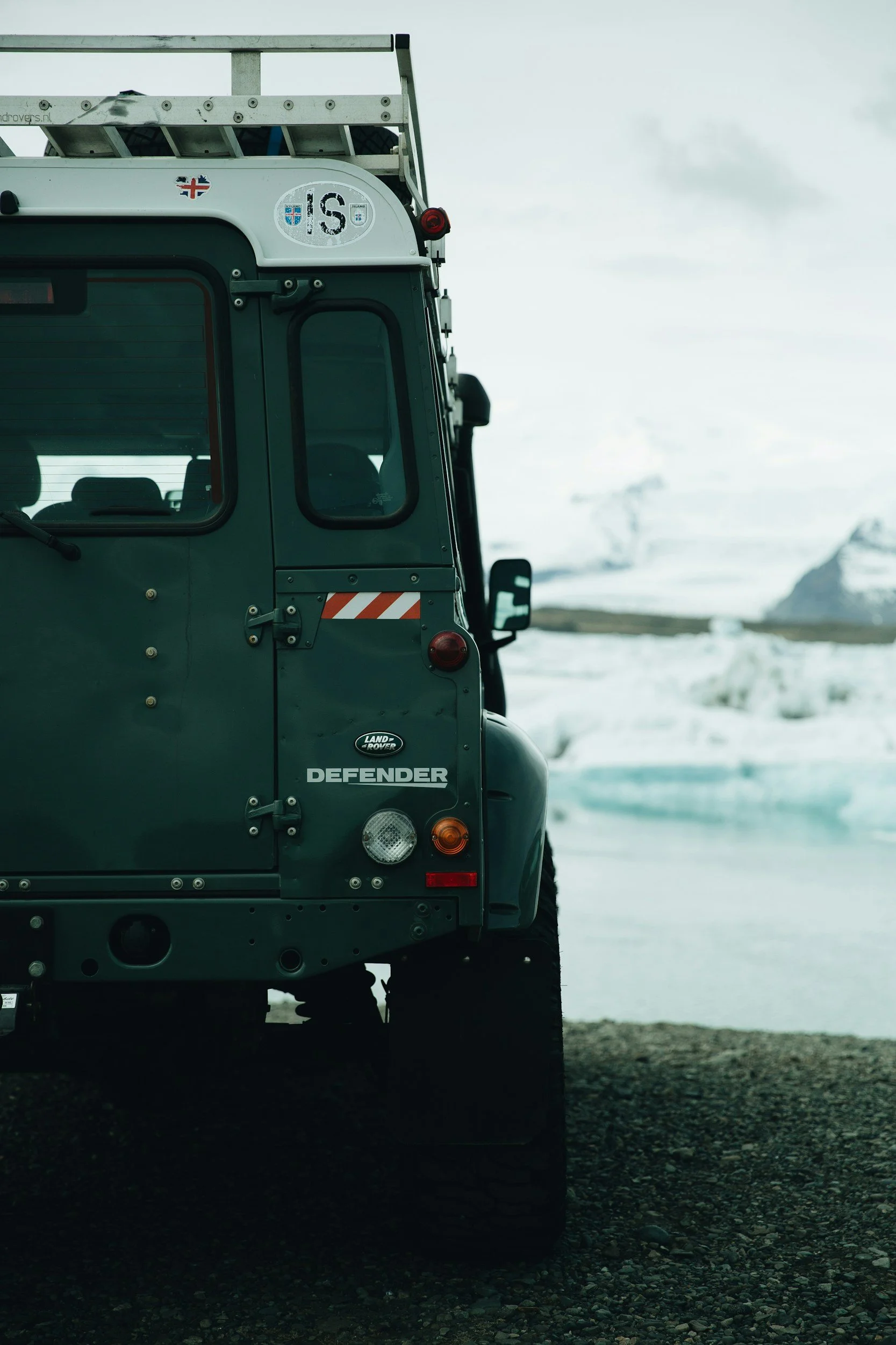 Close-up of the back of a Land Rover Defender vehicle parked on a gravel surface with snowy landscape and mountains in the background.