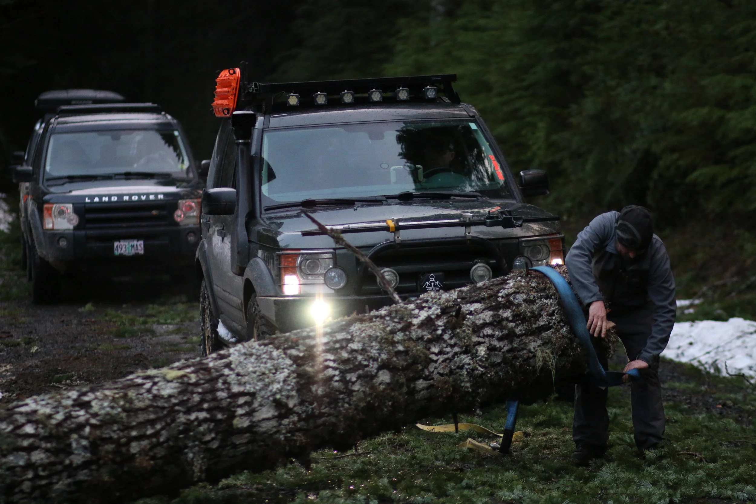Two Land Rover off-road vehicles, a black Land Rover and a second vehicle, are parked in a forested area on a dark, rainy day. A person is working near a fallen tree, possibly clearing or inspecting it.