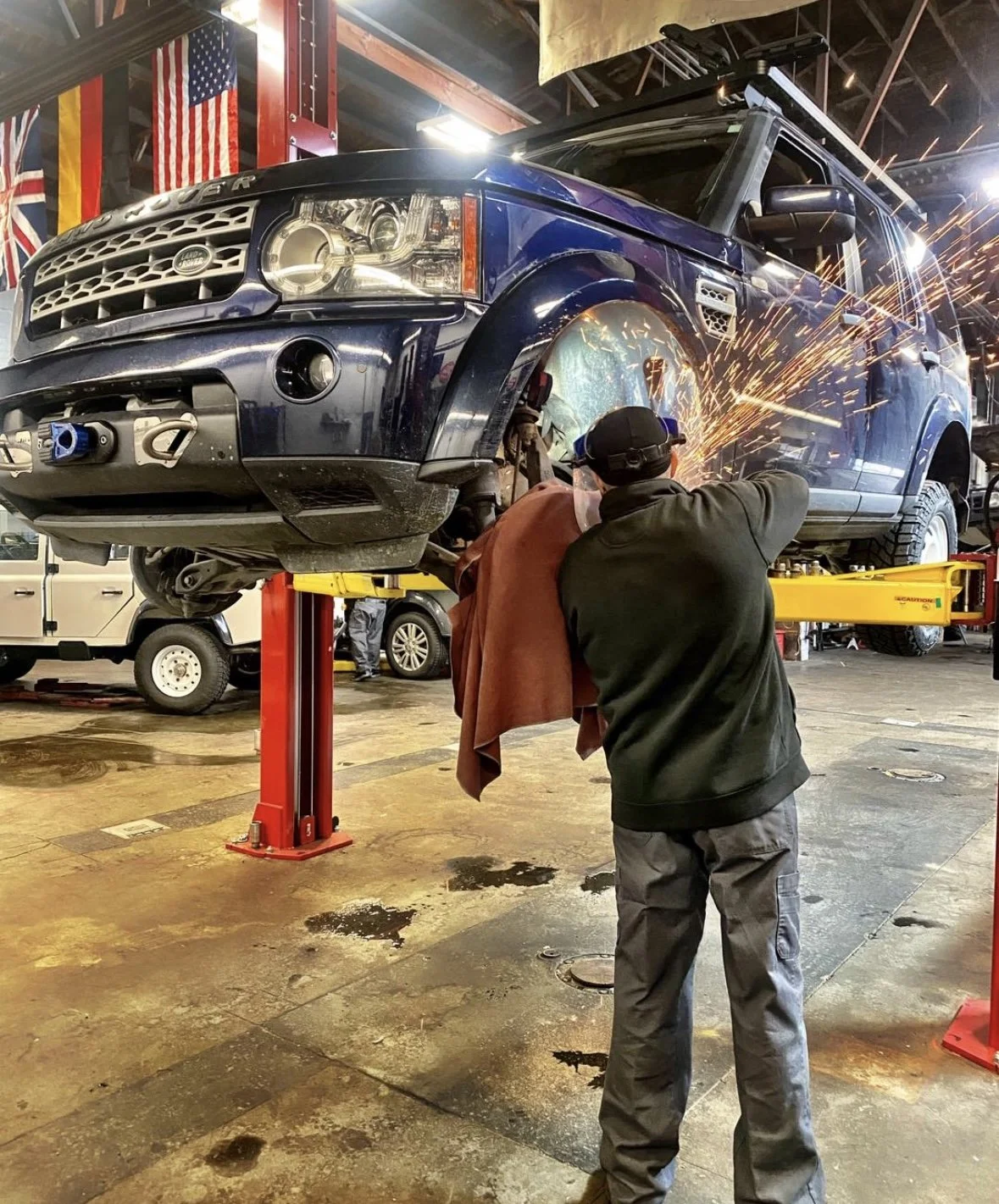 A worker is welding the front wheel area of a blue Land Rover vehicle that is elevated on a hydraulic lift inside an auto repair shop, with sparks flying from the welding process.