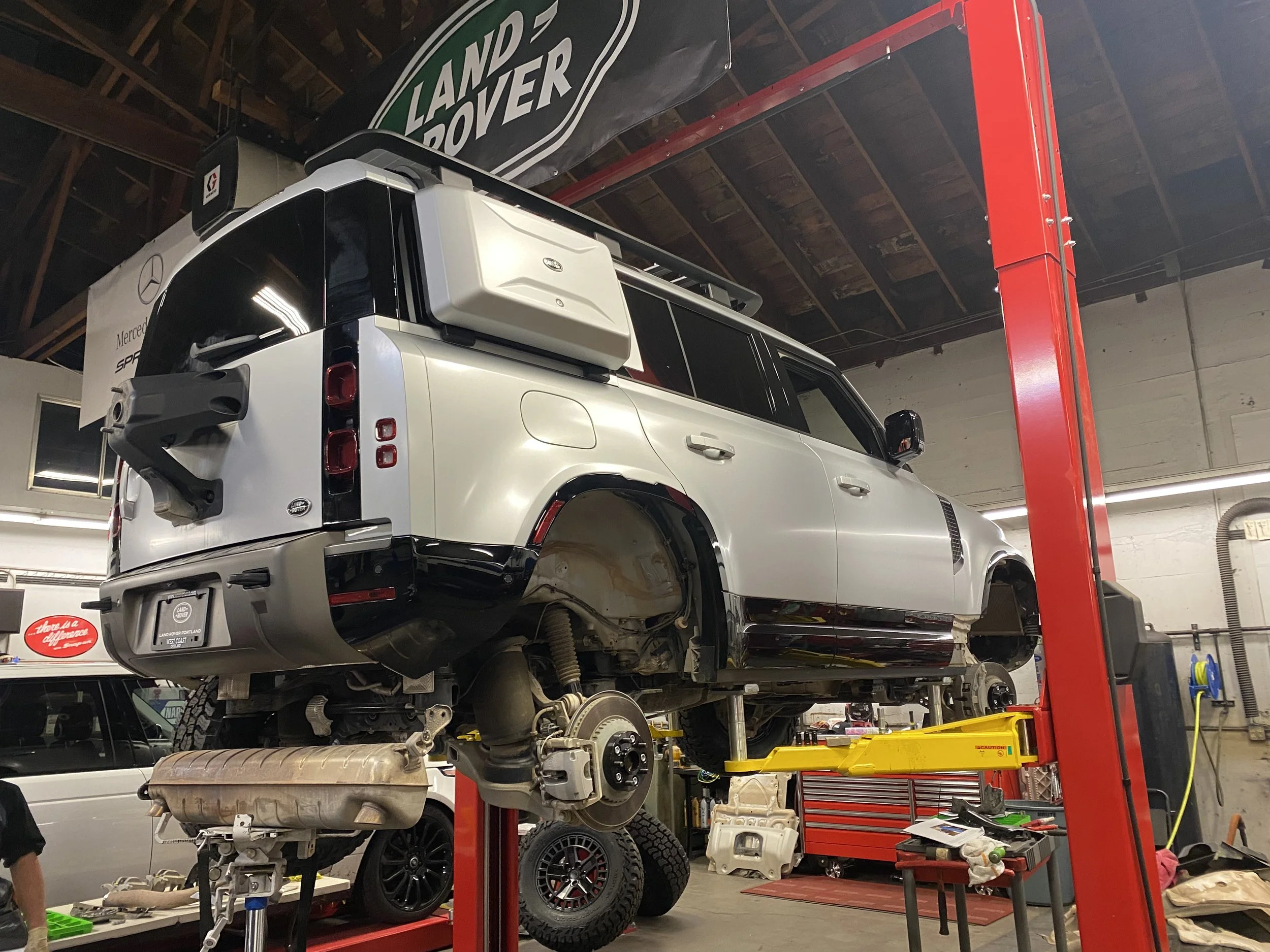 A white Land Rover Defender is lifted on a hydraulic lift in an auto repair shop with tools, tires, and car parts visible around.