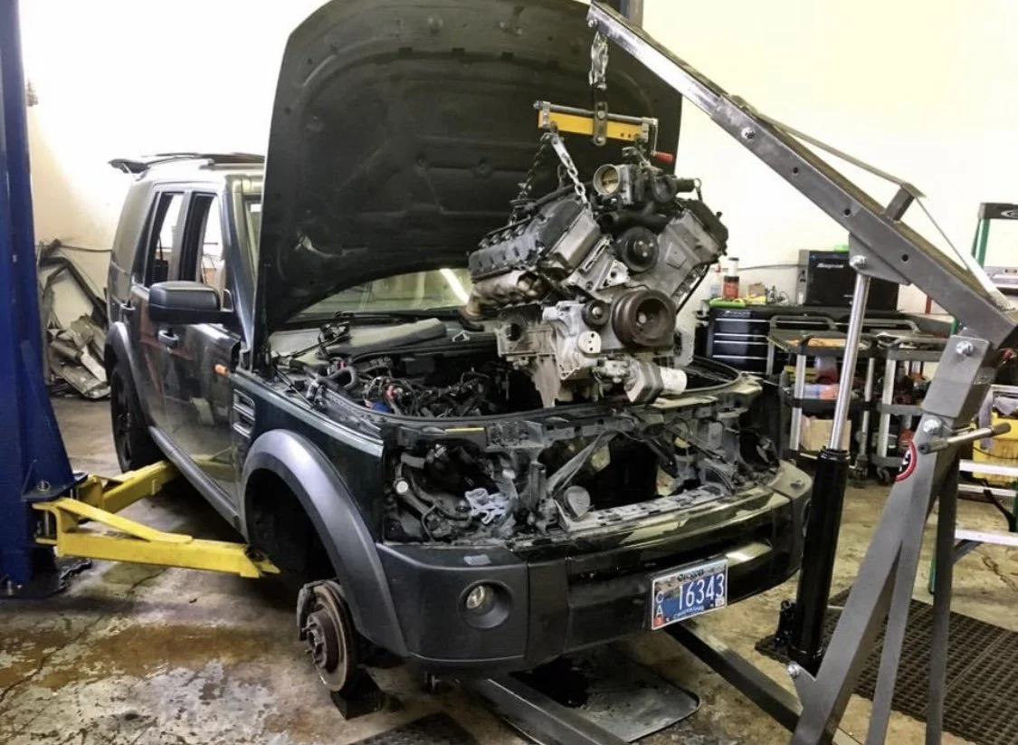 A black Land Rover SUV on a lift in a workshop with its hood open and an engine hoisted above the engine bay, indicating engine repair or replacement.