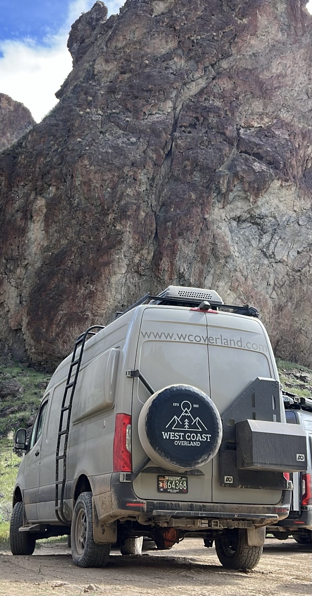 A tan Sprinter van parked in front of a large rocky mountain with a partly cloudy sky.