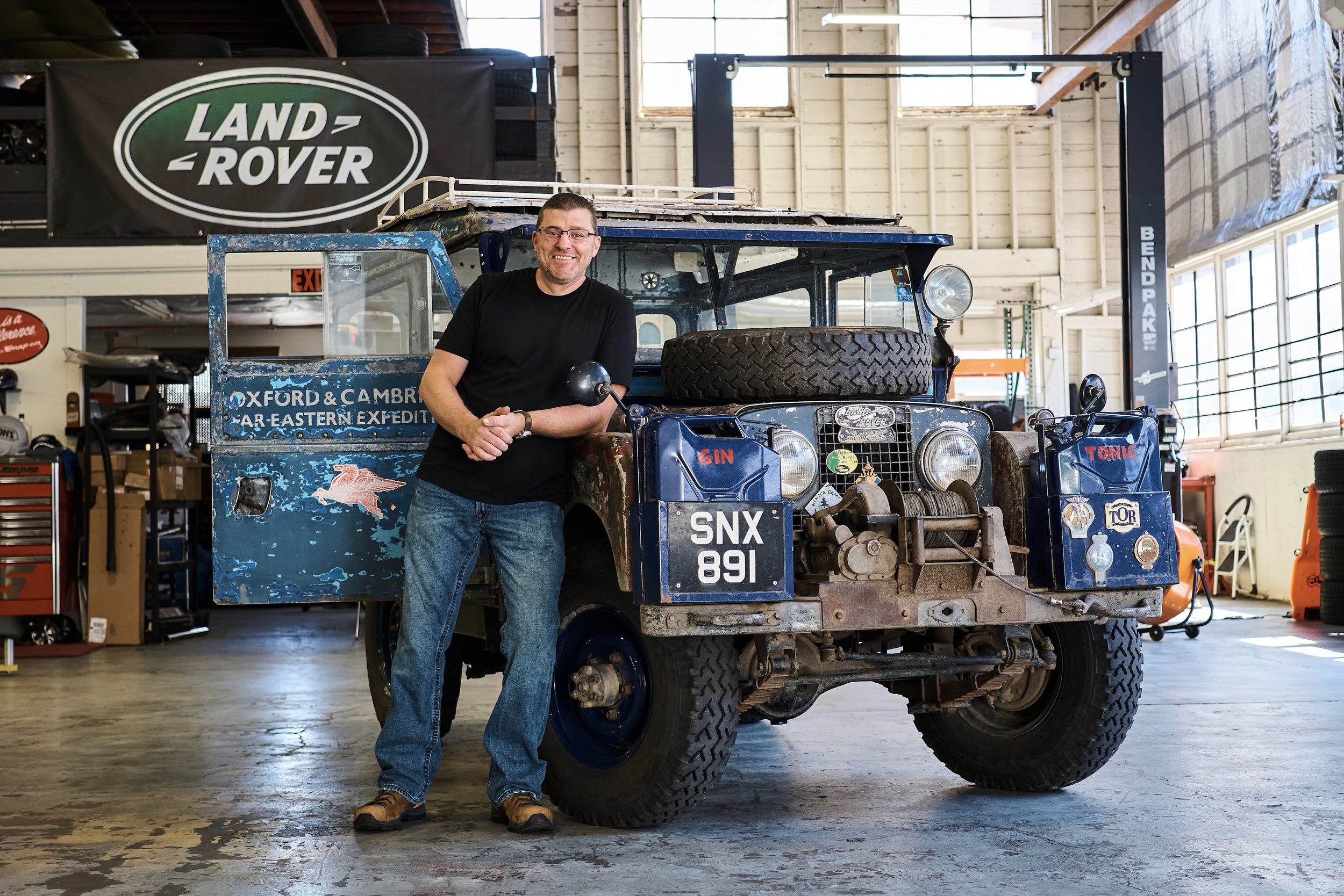 A man smiling and standing inside a workshop next to an old, partially disassembled Land Rover vehicle. The workshop has a large Land Rover banner on the wall, toolboxes, and various equipment.