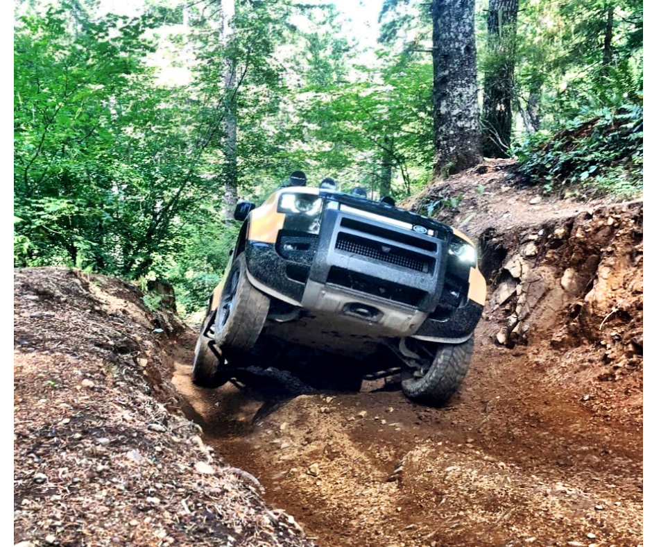 A yellow and black off-road Land Rover Defender 110 navigating a rugged dirt trail in a forested area with tall green trees.