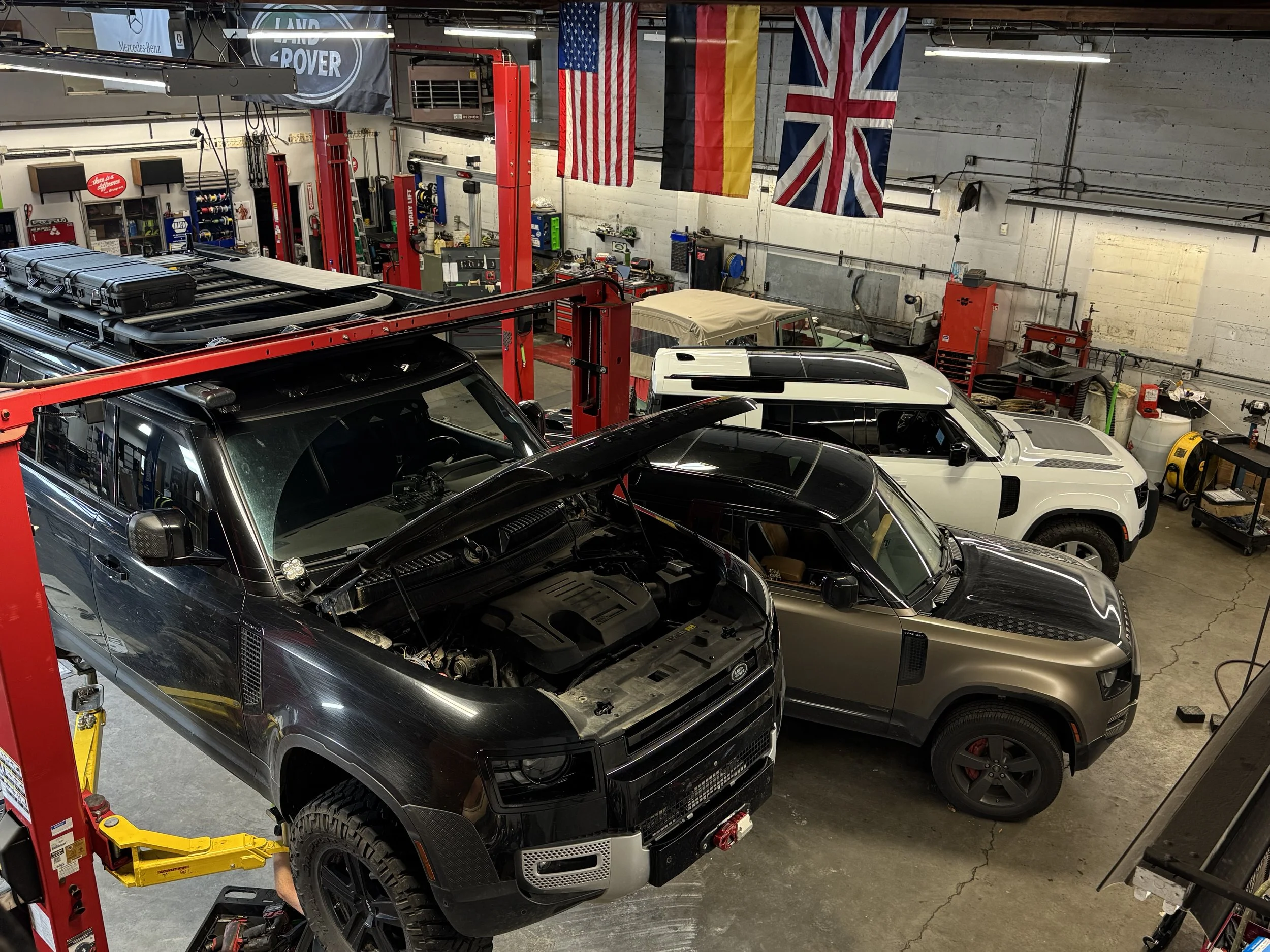 An automotive workshop with a black SUV on a hydraulic lift in front of multiple vehicles including a white car and a beige vehicle. The wall displays American, German, and British flags, with various tools, equipment, and automotive supplies organiz