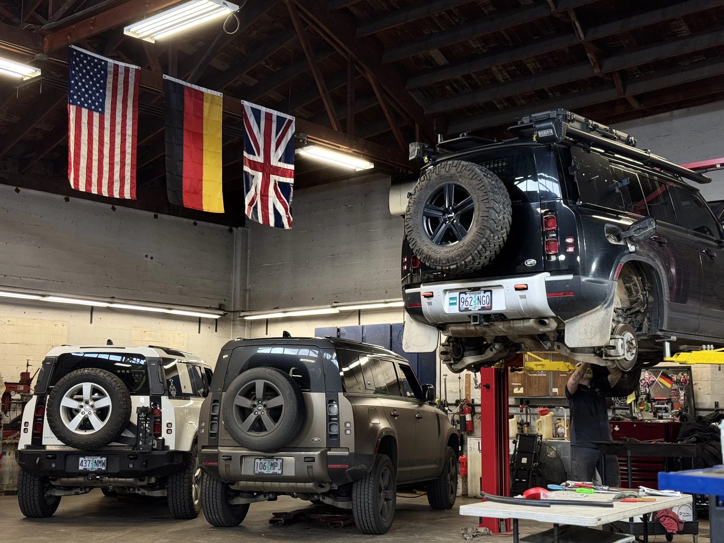 Inside an auto repair shop with Land Rover Defenders on lifts. Flags of the United States, Germany, and the United Kingdom hang from the ceiling. A mechanic is working on a black Defender 110. 