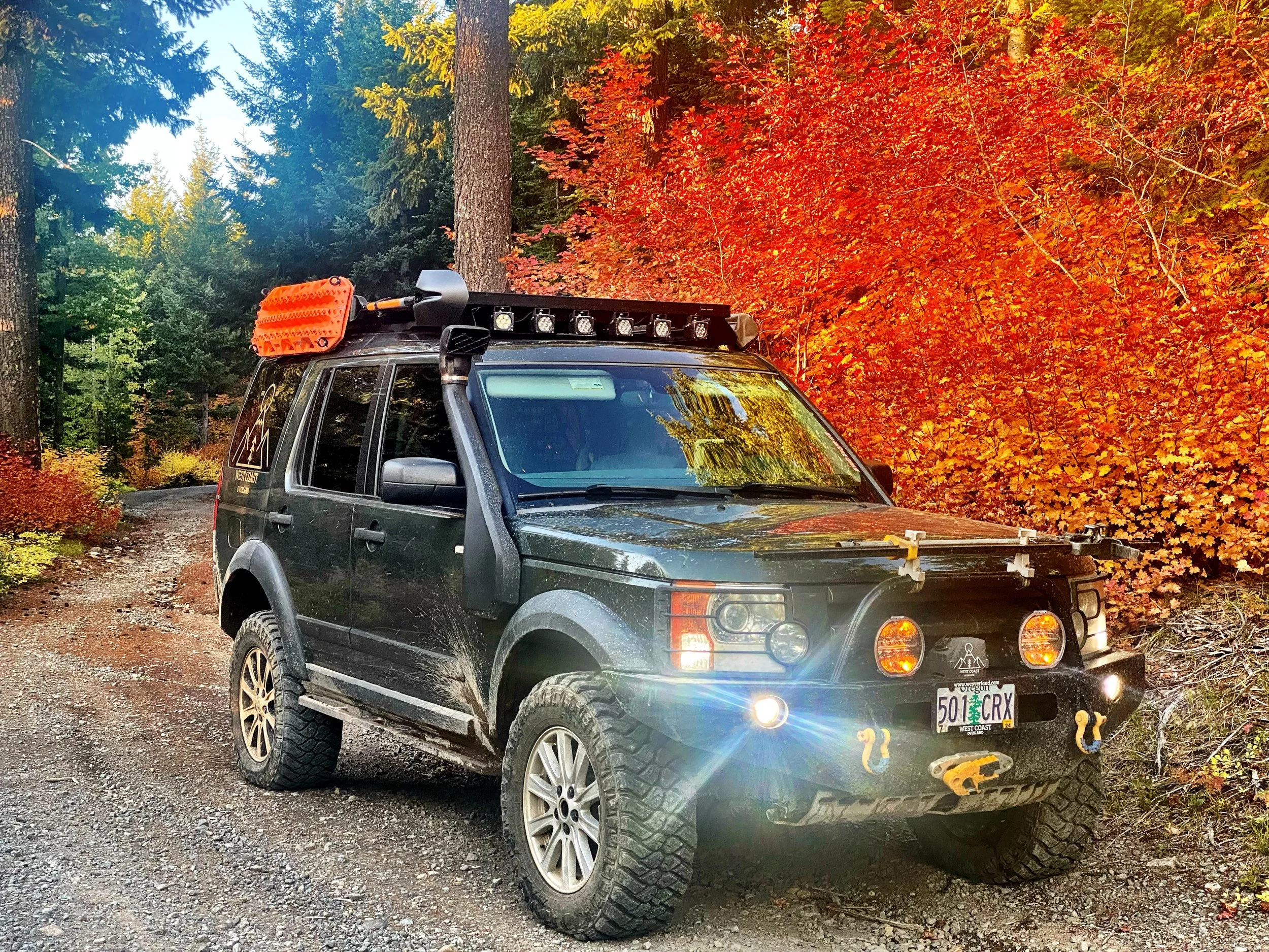 A greej off-road Land Rover LR3 parked on a dirt trail in a forest during fall, surrounded by trees with red, orange, and green leaves. The vehicle has equipment on the roof, including lights, a roof rack, and a large orange snow track.