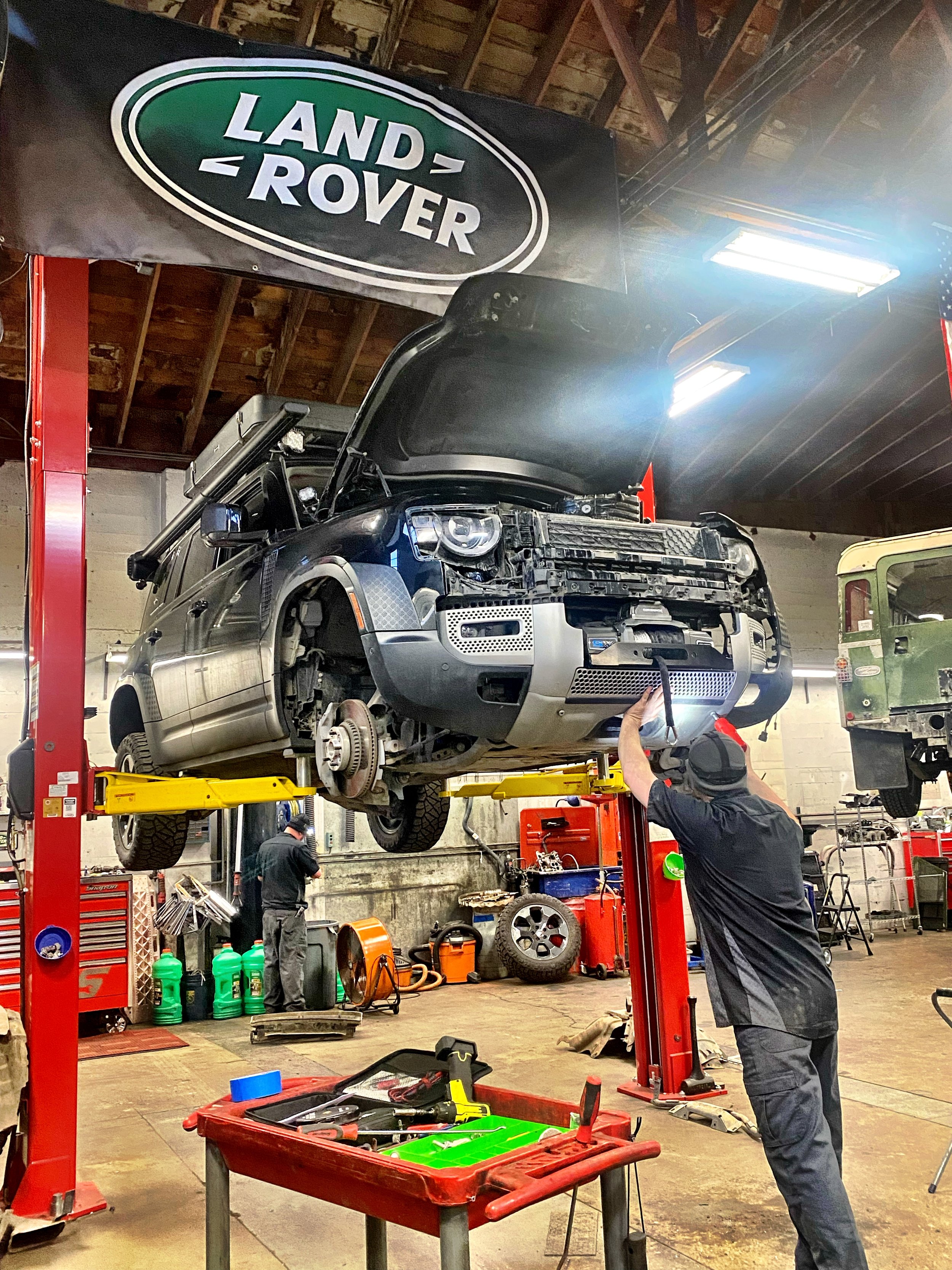 A mechanic working on a Land Rover vehicle that is elevated on a hydraulic lift inside an automotive repair shop. The vehicle's front bumper is removed, and the mechanic is using a tool underneath it. The shop has various tools and equipment, and a L