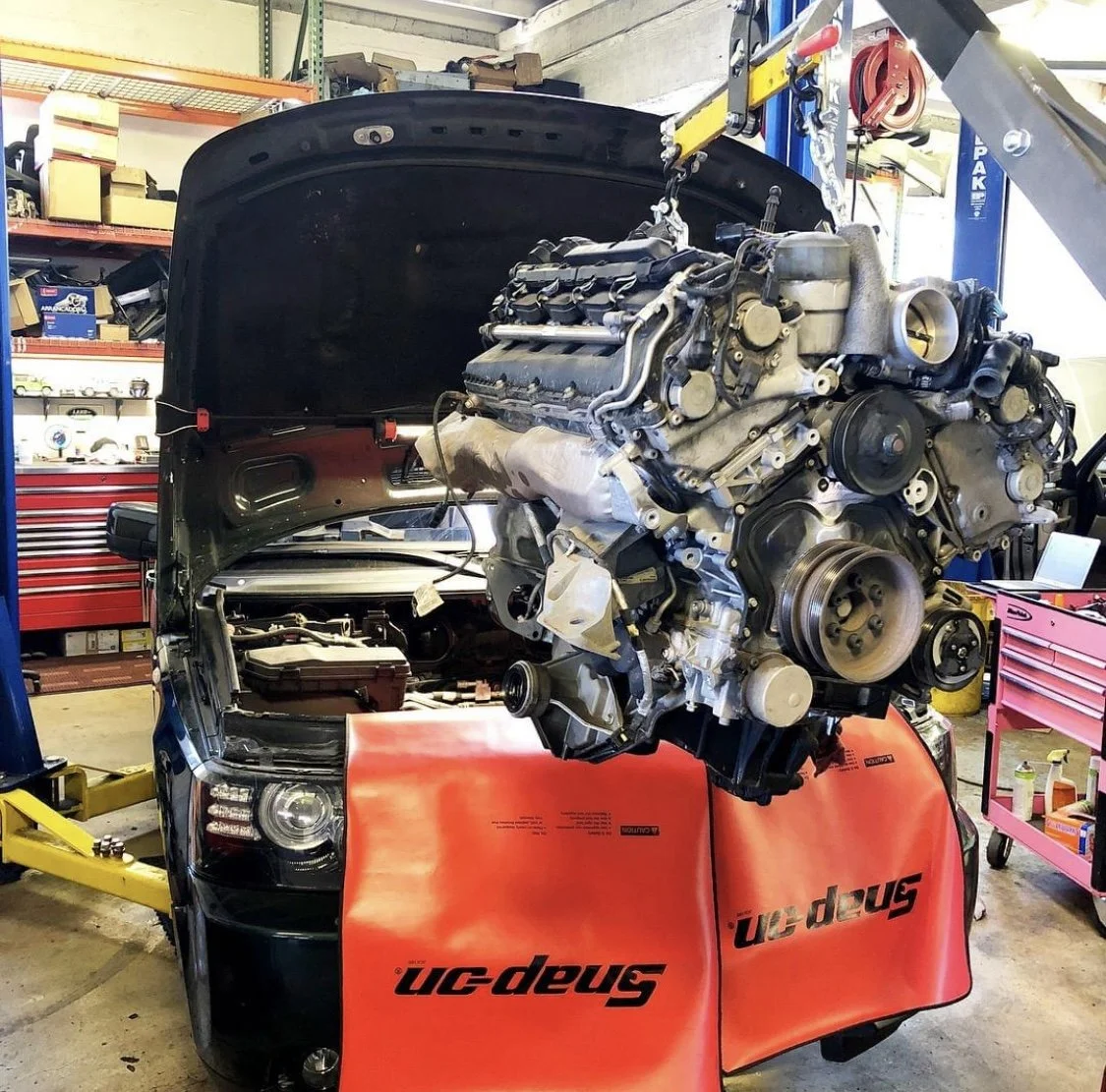A Land Rover repair shop with a lifted car engine being installed into a black car with the hood open. The engine is hanging from a hoist, and the shop has various tools and equipment around.
