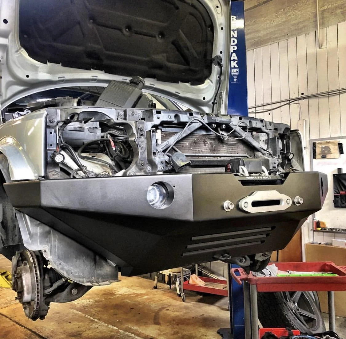 Front of a Land Rover with the hood open, showing the engine bay with front bumper removed, in a garage or workshop.