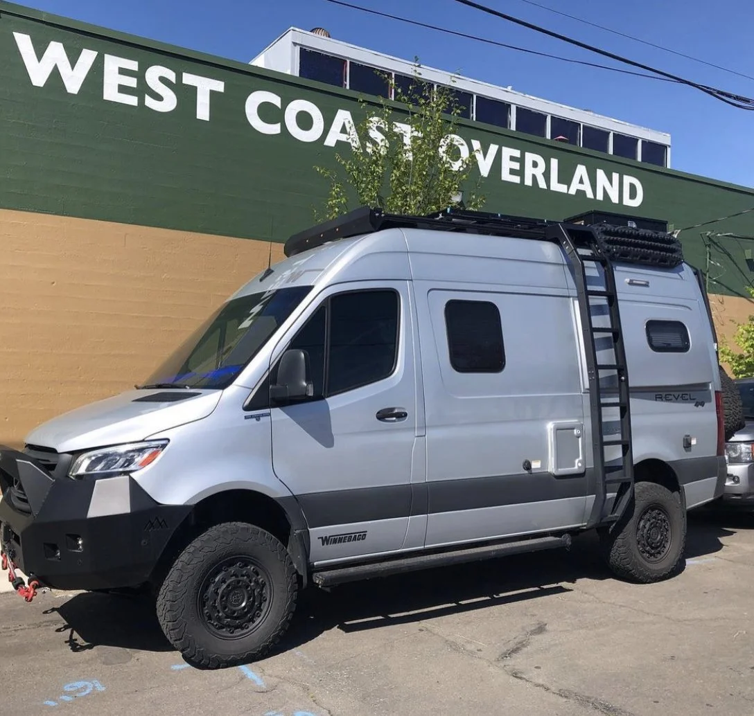 A silver off-road Sprinter van parked in front of a building with a green wall that says 'West Coast Overland.'