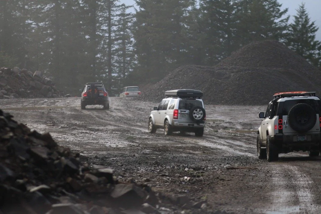 Four Land Rover SUVs driving on a muddy, gravel road through a forested area, with tall trees and large piles of dirt in the background.