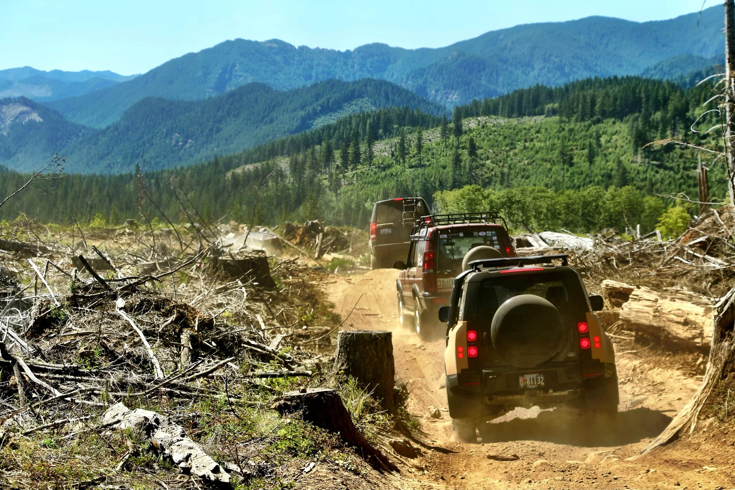 Three off-road vehicles driving on a dirt trail through a forested mountain area, with fallen trees and debris on the ground.