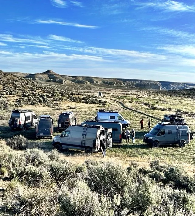 A group of Sprinter vans parked in a desert landscape with low shrubs, under a blue sky with scattered clouds.