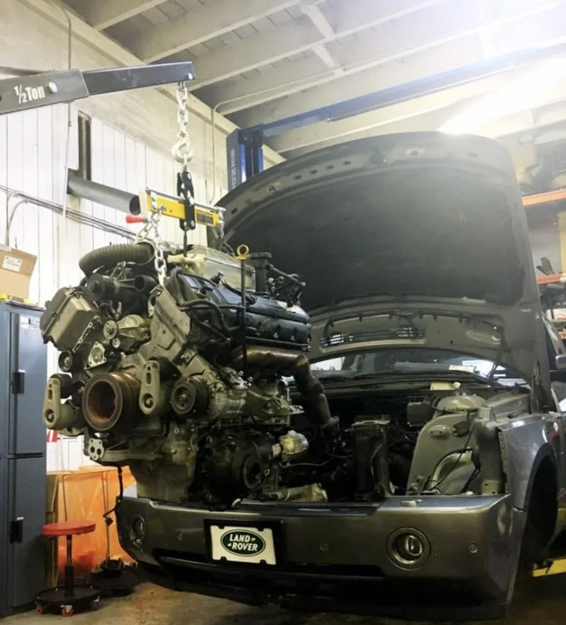 A Land Rover vehicle with its front end and engine removed during repair inside a garage. The engine is being lifted out by a hoist.