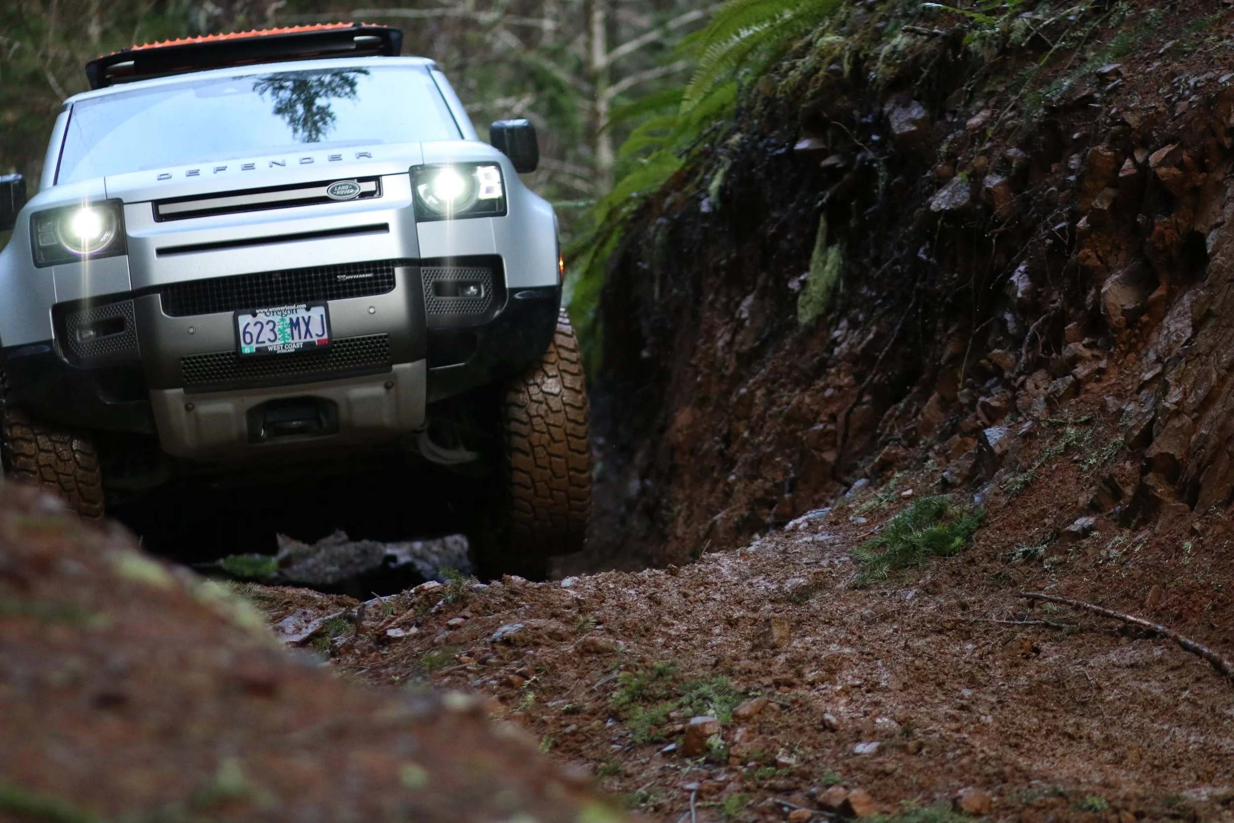 A silver Land Rover Defender climbing a muddy trail in a forested area, with dense greenery and rocks beside the path.