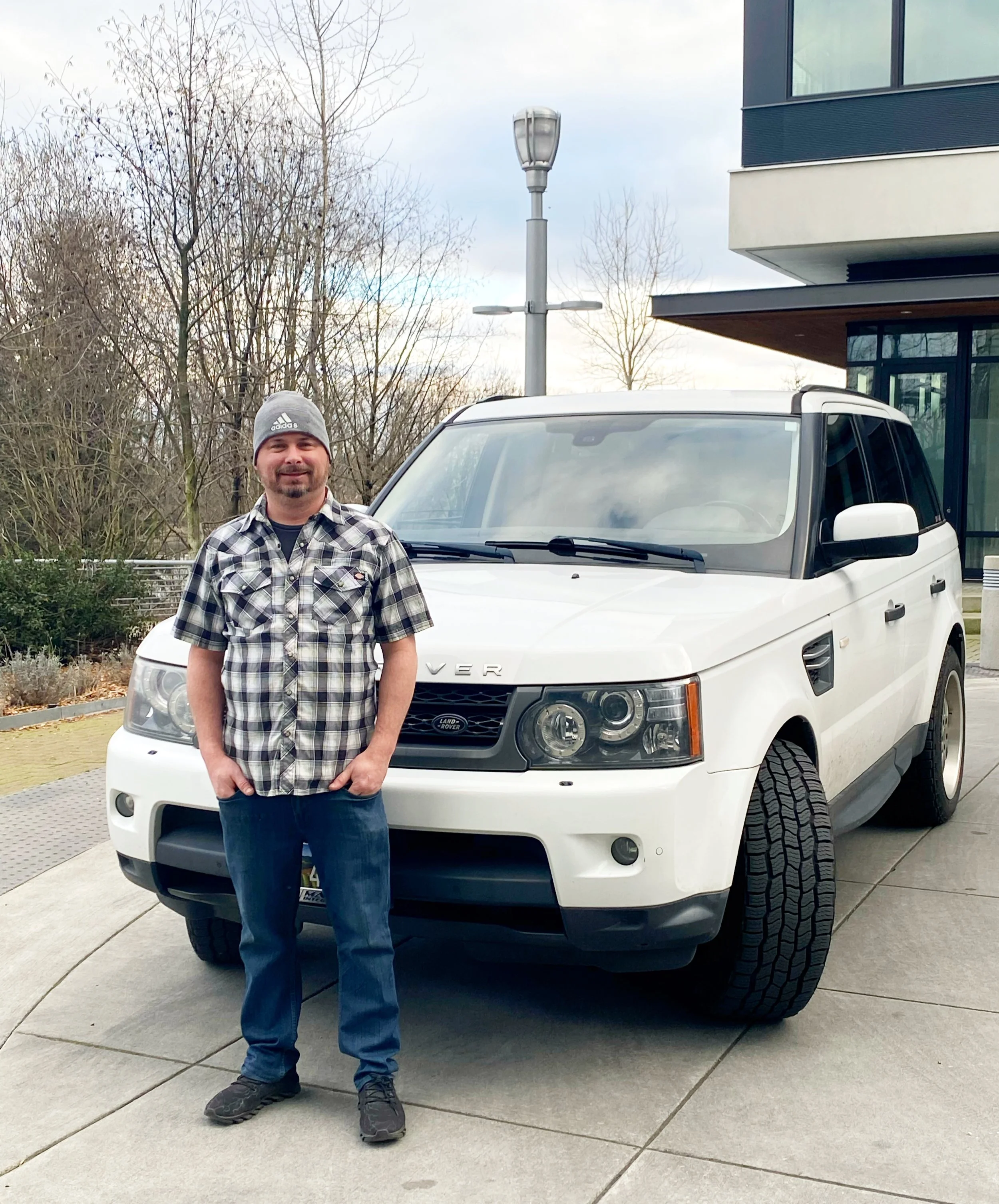 A man wearing a gray beanie, a plaid shirt, and jeans stands in front of a white Land Rover SUV on a concrete driveway. Behind him are leafless trees, a streetlight, and a modern building with large windows.