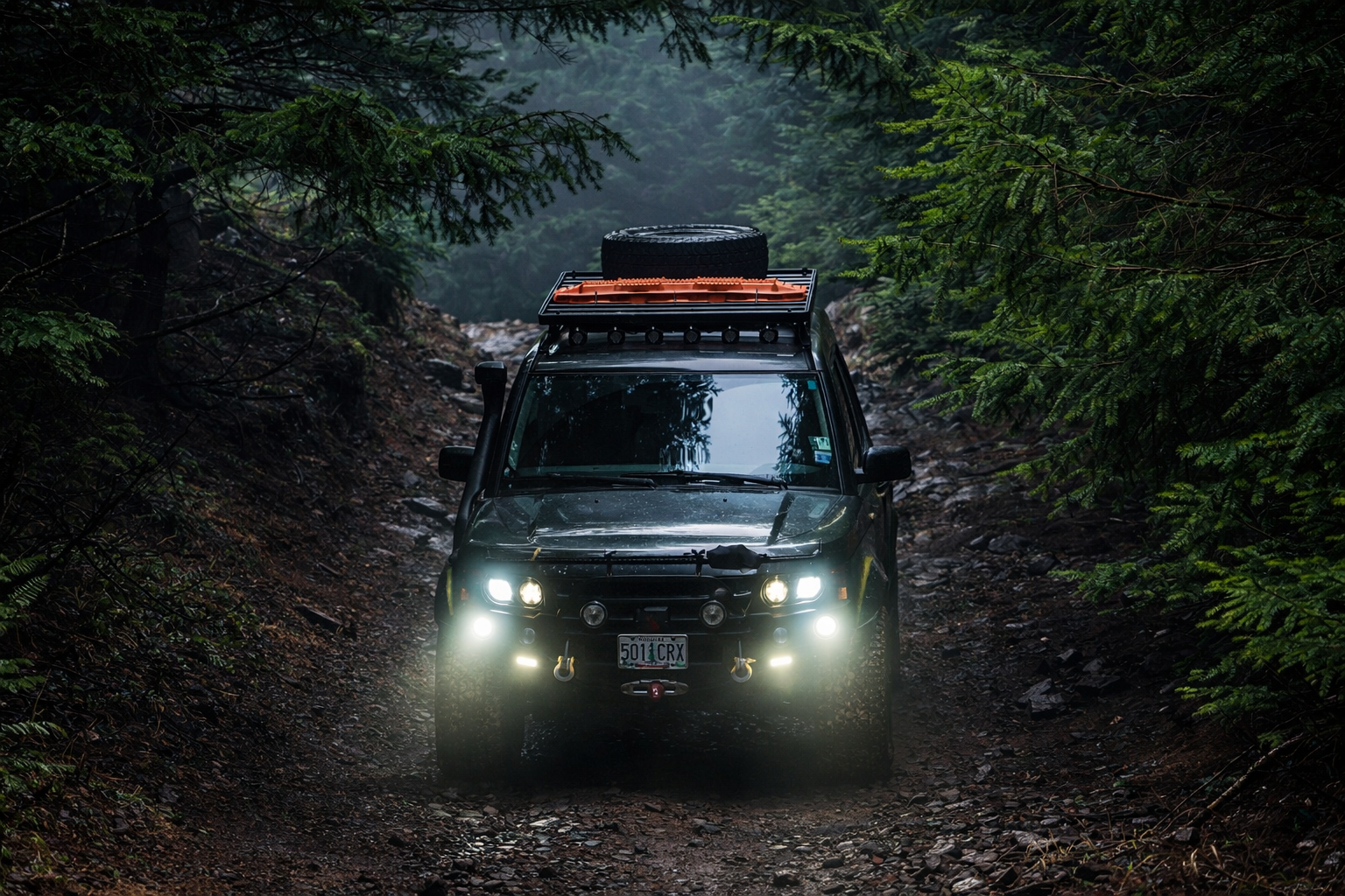 A green Land Rover LR3 driving through a dense forest on a rough, rocky trail with its headlights and fog lights on, carrying a spare tire on a roof rack.