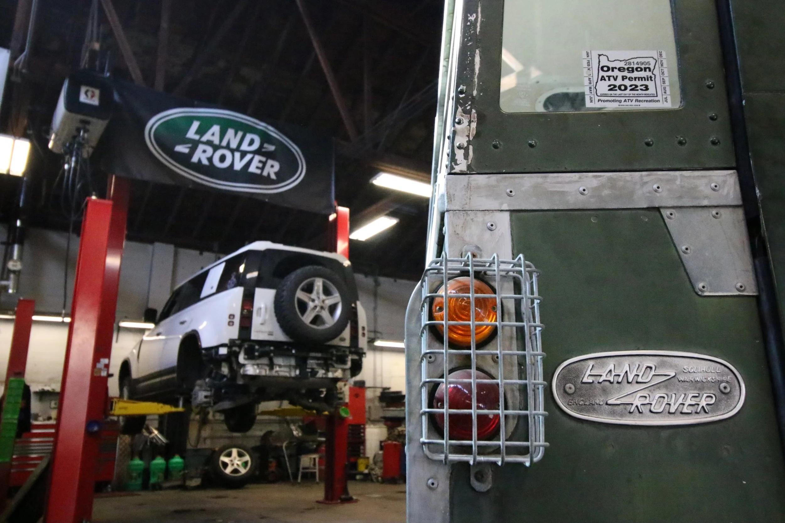 Inside an auto repair shop, a white Land Rover is elevated on a hydraulic lift with its underside visible. A large black banner with the Land Rover logo hangs in the background. To the right, a metal box with an Oregon ATV permit sticker is attached 