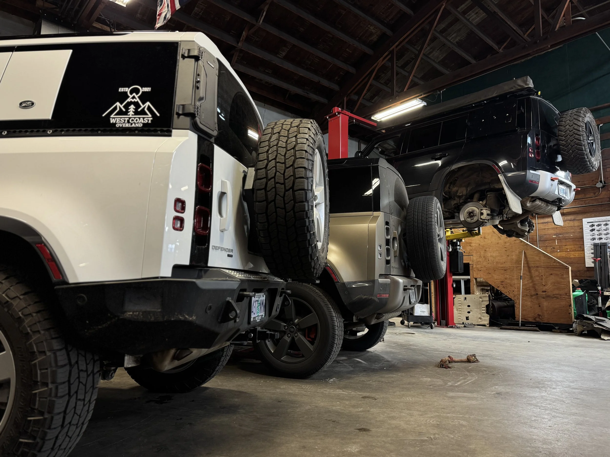 Multiple vehicles, including a white and a black SUV, are parked inside a garage or workshop with a wooden ceiling and automotive tools and equipment visible.