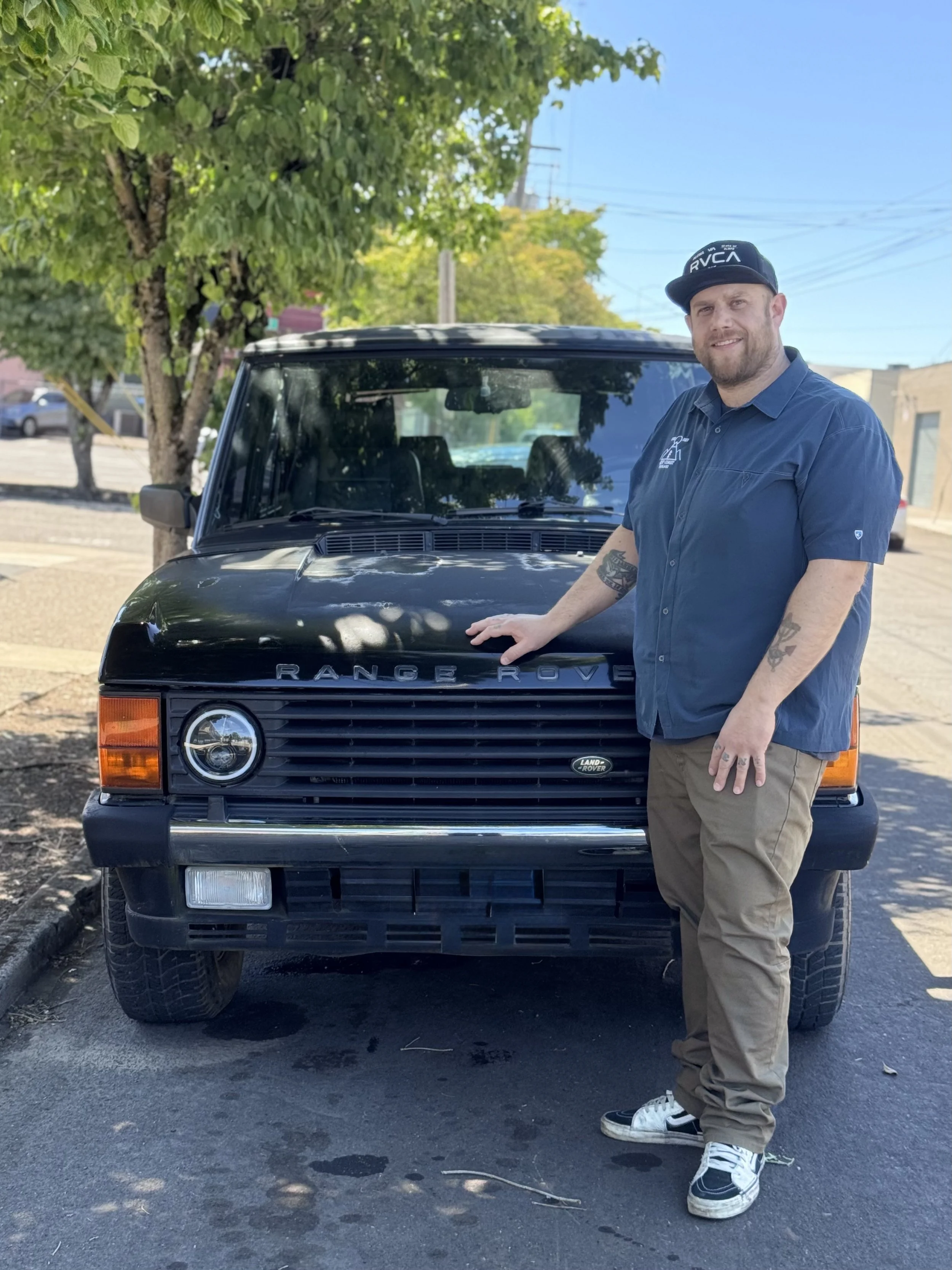 A man standing next to a black Land Rover Range Rover vehicle in an outdoor parking lot on a sunny day. He is wearing a dark blue shirt, khaki pants, and sneakers, and is smiling at the camera.