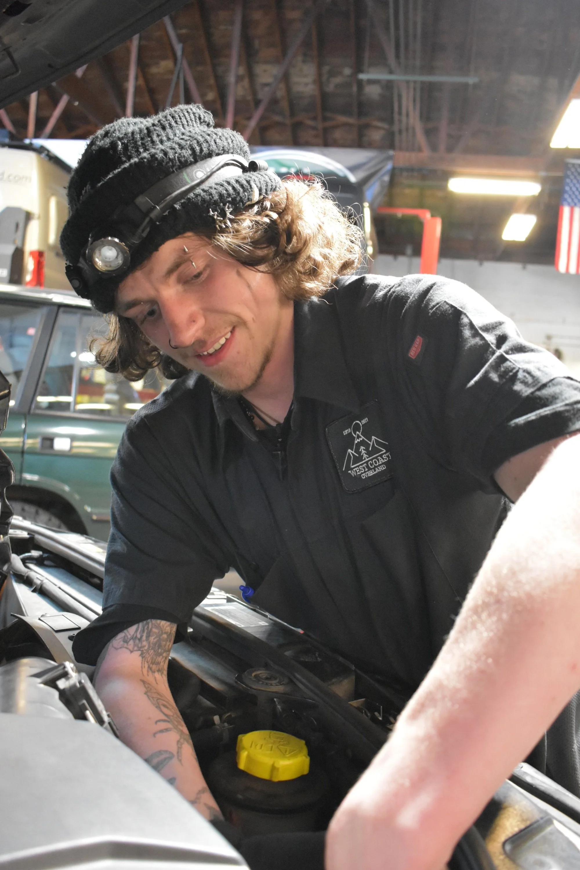 A young man with tattoos on his arm working on a car engine inside a garage, wearing a black beanie, headphones, and a black jacket with a logo that reads 'West Coast Outlawz'.
