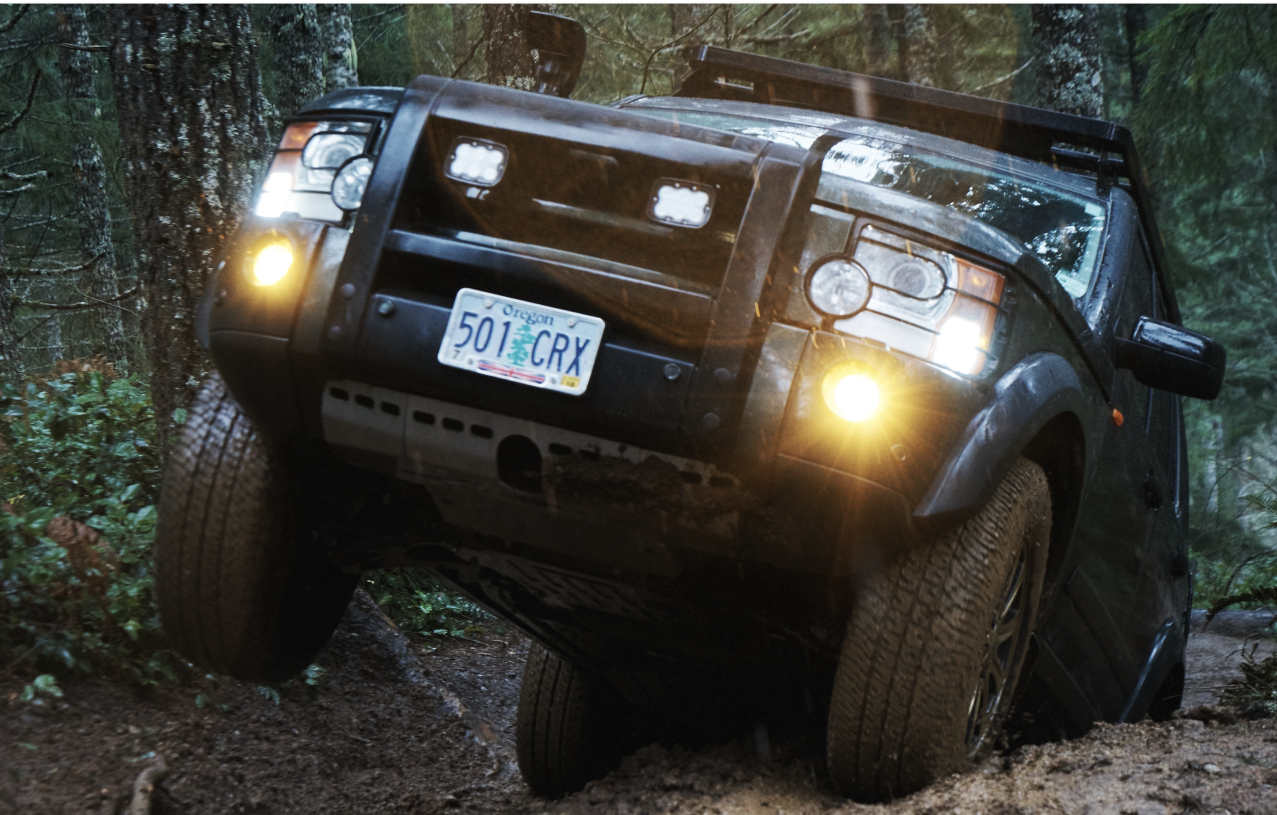 A black off-road Land Rover SUV with bright headlights and Oregon license plate partially tilted on uneven dirt terrain surrounded by dense green forest.