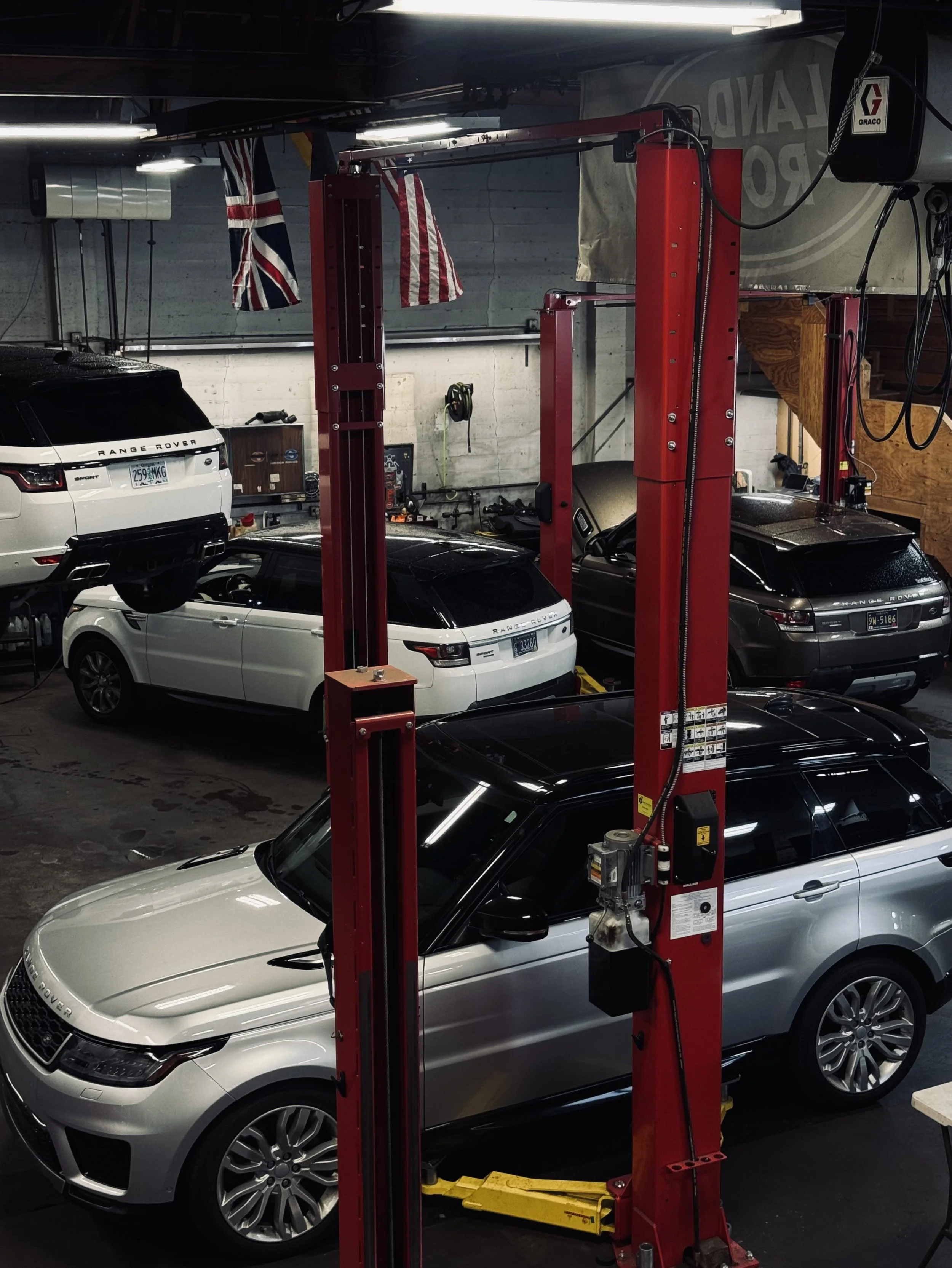 Multiple Range Rover vehicles parked inside a garage with a red automotive lift in the foreground. A Union Jack flag hangs on the wall on the left side of the image.